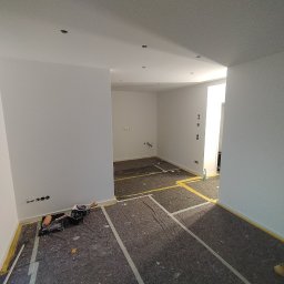 USŁUGI BUDOWLANE Oktawian Rożenko - Wnętrze po malowaniu na biało, zabezpieczona podłoga, widoczne narzędzia malarskie i drabina.