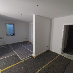 USŁUGI BUDOWLANE Oktawian Rożenko - Przygotowane do malowania wnętrze z zabezpieczoną podłogą, świeżo pomalowane ściany na biało, widoczna drabina malarska i fragment okna.