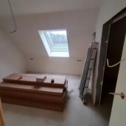 Poddasze w trakcie prac wykończeniowych: przygotowane panele podłogowe w oryginalnych opakowaniach, aluminiowa drabina oparta o ścianę, okno dachowe z widokiem na drzewa.