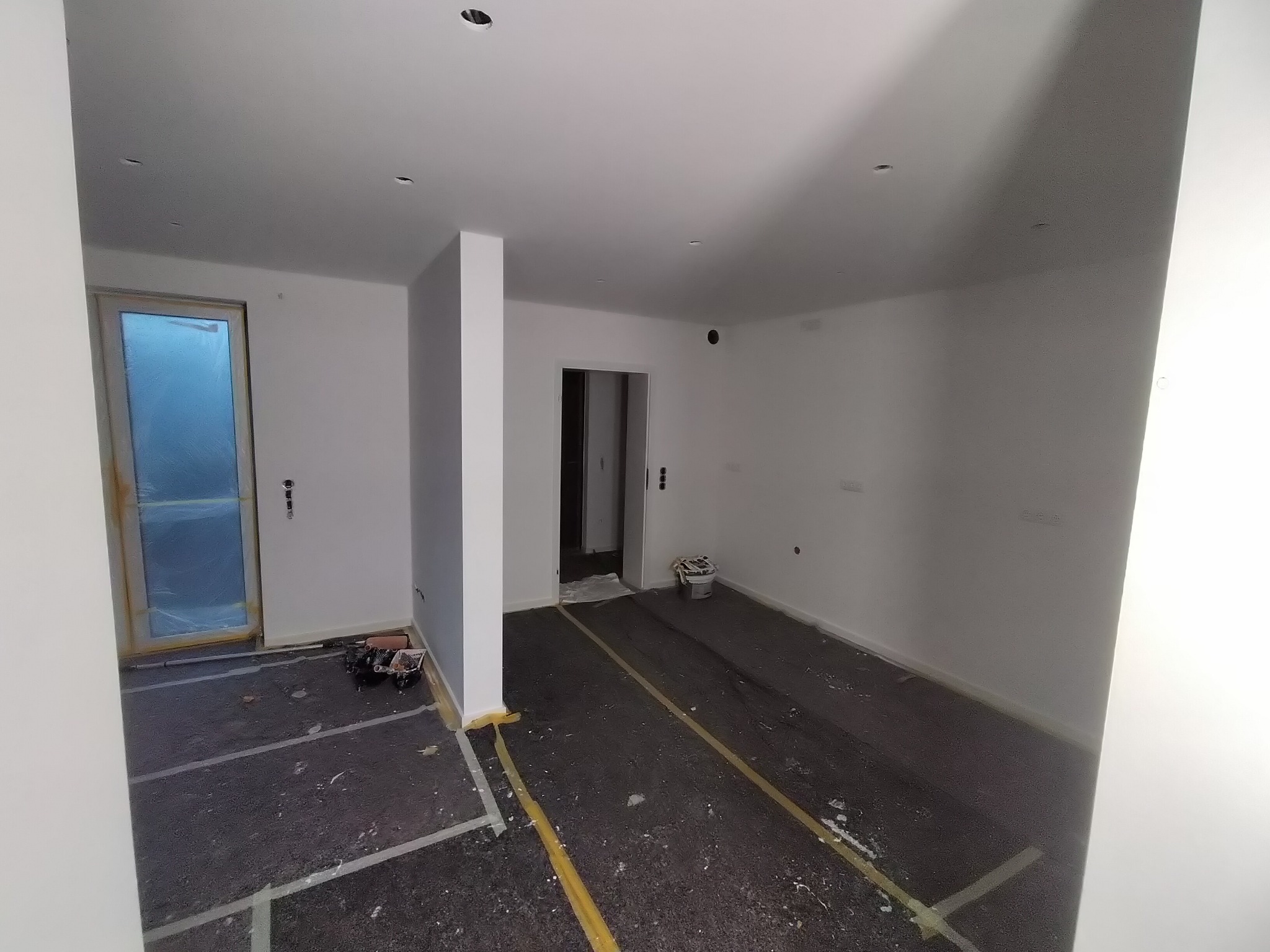 Przygotowane do malowania wnętrze: zabezpieczona folią podłoga, oklejone listwy przypodłogowe, widoczne narzędzia malarskie przy drzwiach, surowe białe ściany.