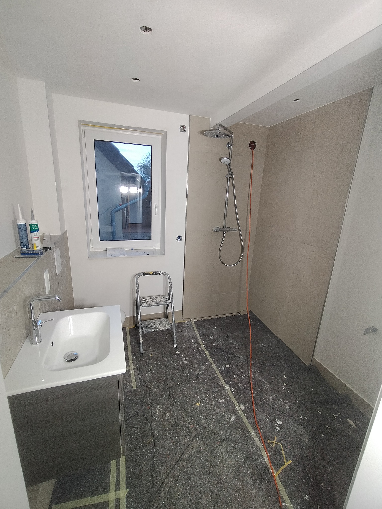 Łazienka w trakcie wykańczania: zamontowana umywalka z szafką, kabina prysznicowa z deszczownicą, drabinka malarska, podłoga zabezpieczona folią malarską, widoczne silikony i materiały budowlane.
