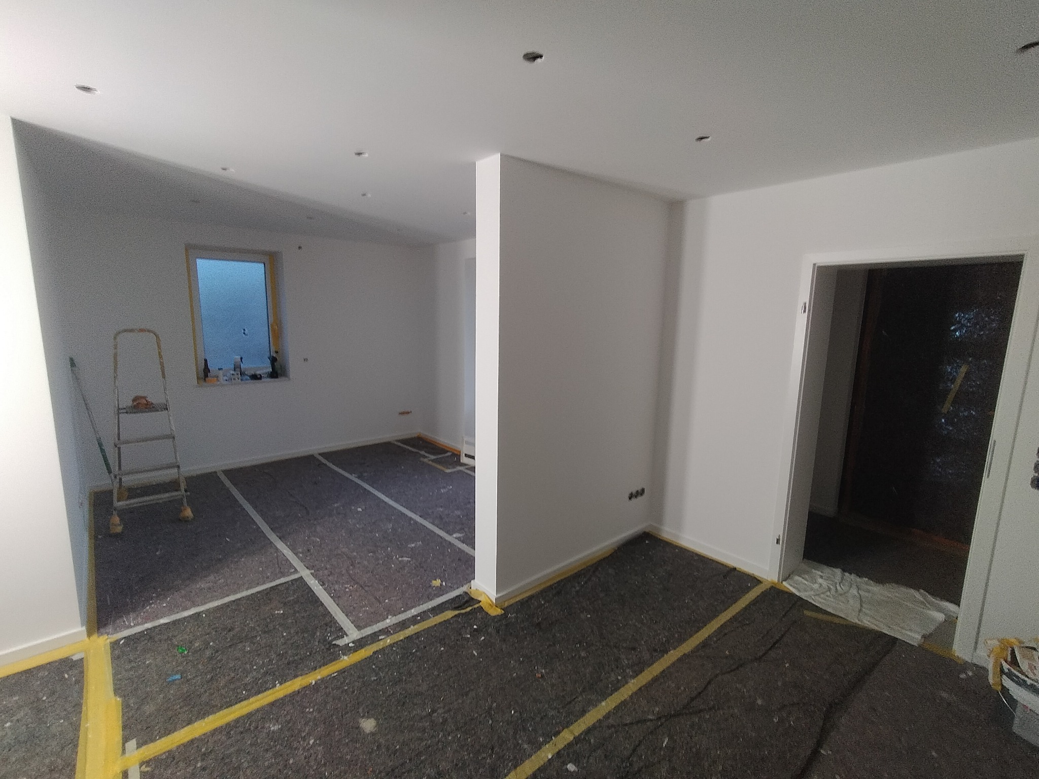 Przygotowane do malowania wnętrze z zabezpieczoną podłogą, świeżo pomalowane ściany na biało, widoczna drabina malarska i fragment okna.