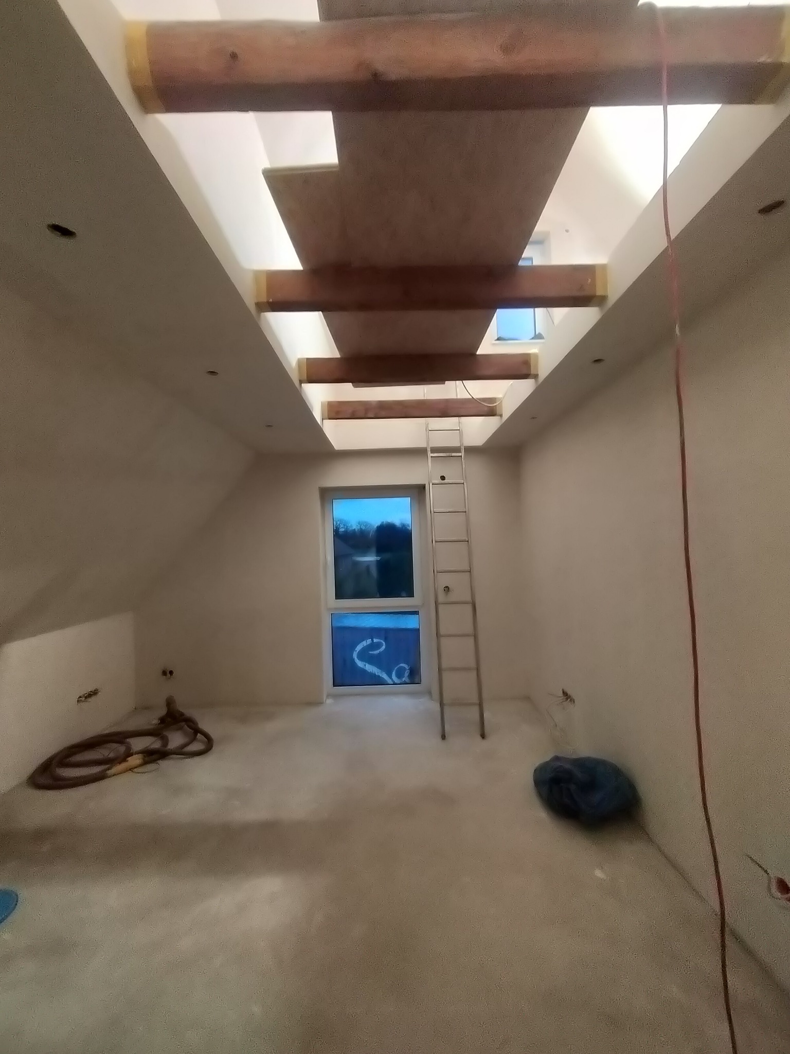 Poddasze w trakcie adaptacji: widoczne drewniane belki stropowe, surowa wylewka, okno dachowe i drabina oparta o ścianę. Przygotowanie do wykończenia wnętrza.