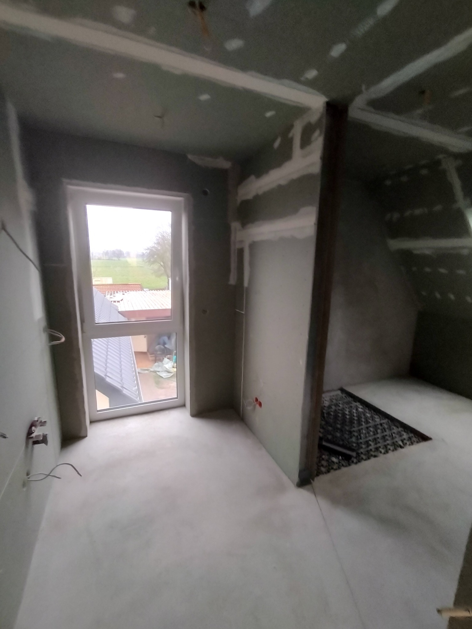 Surowe wnętrze w trakcie prac wykończeniowych: widoczne szpachlowane ściany, wylewka podłogowa i okno z widokiem na zewnątrz; instalacja elektryczna wystaje ze ściany.