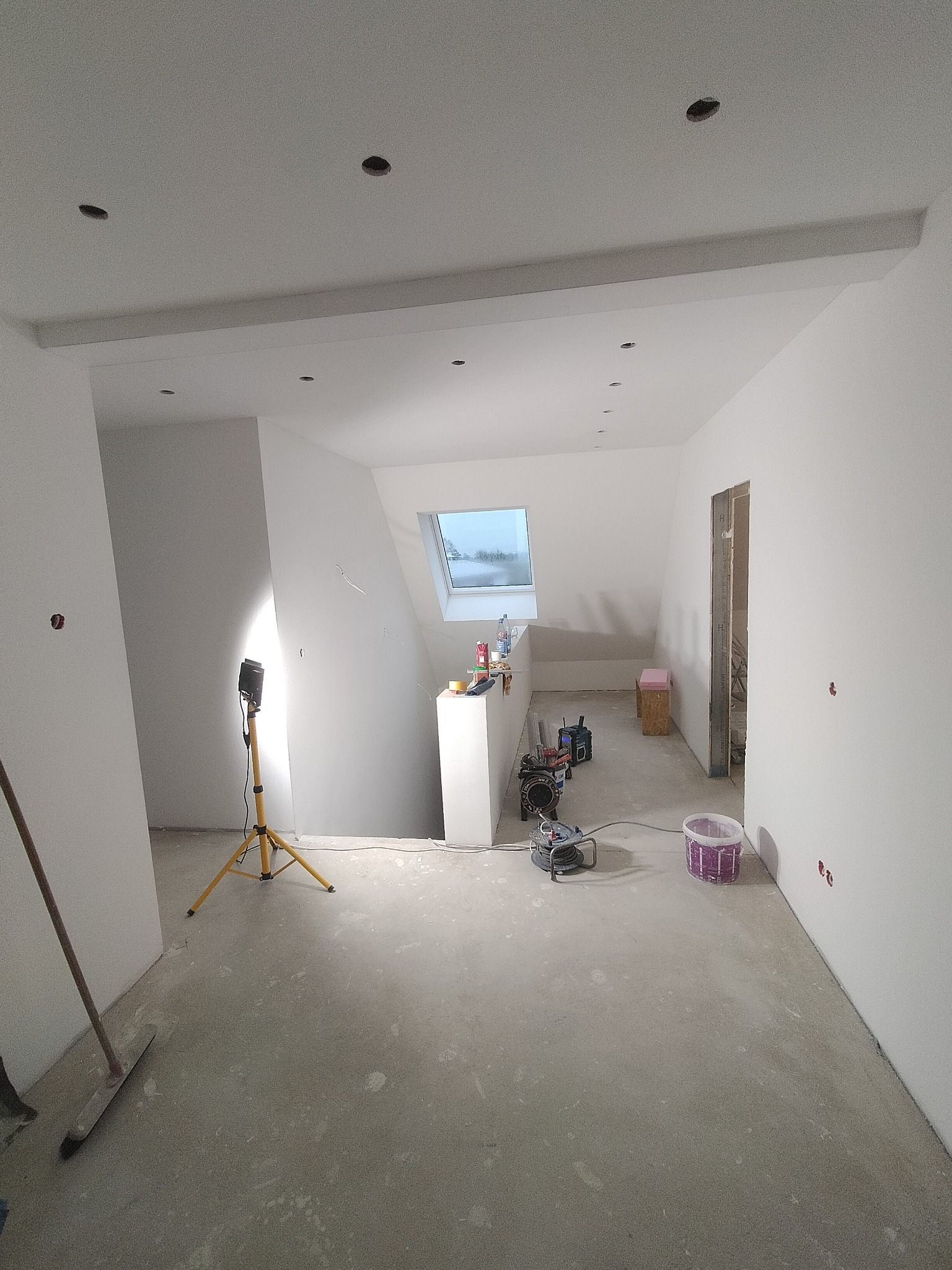 Wnętrze w trakcie prac wykończeniowych: białe ściany, sufit z punktowymi otworami na oświetlenie, podłoga w stanie surowym, widoczna lampa budowlana na statywie i narzędzia.