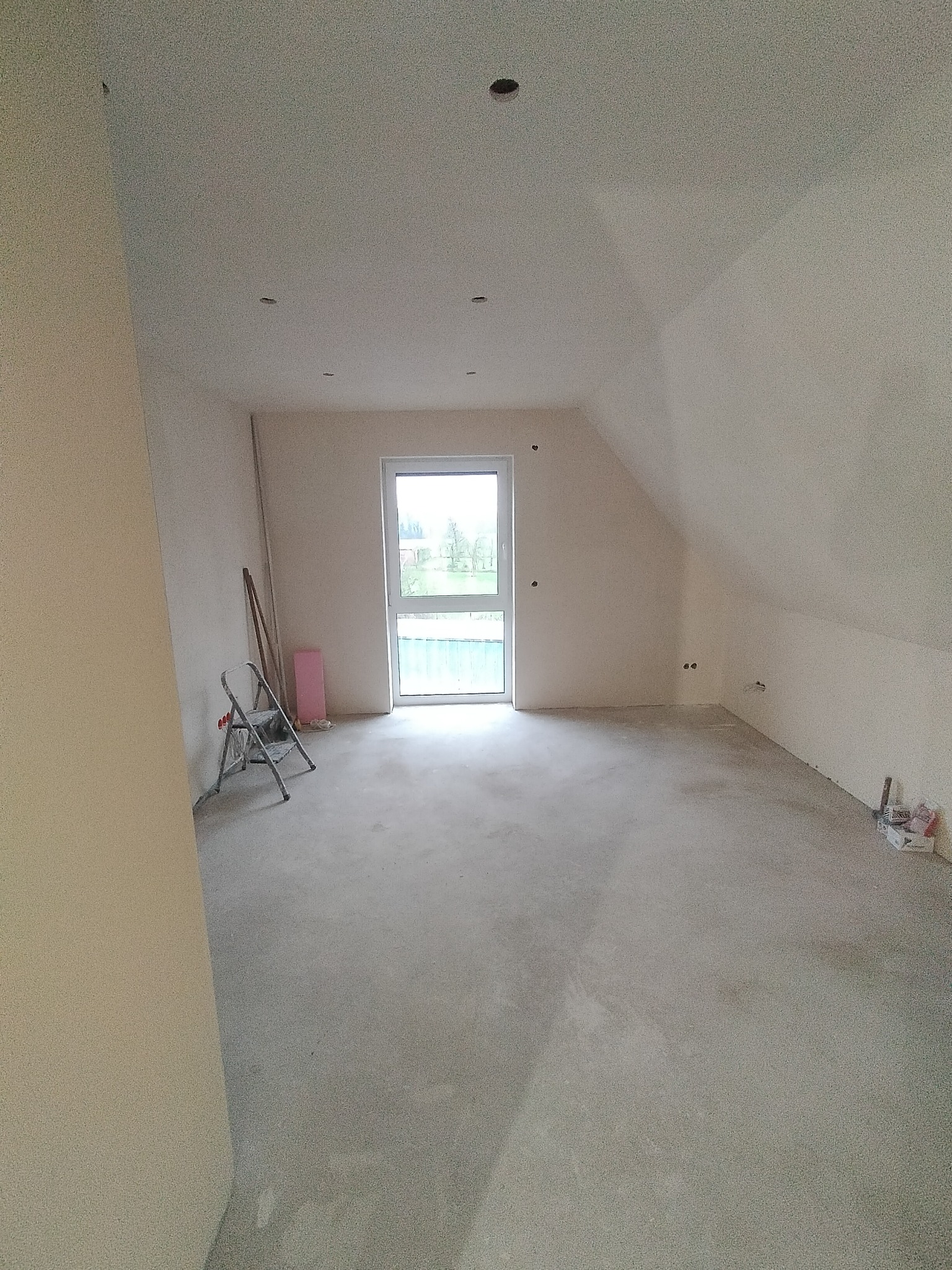 Poddasze w stanie deweloperskim z oknem balkonowym, widoczne ślady po instalacji elektrycznej, drabina malarska i materiały budowlane przy ścianie.