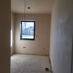 ART-INVEST Damian Solarski - Pokój w trakcie wykańczania: ściany pokryte świeżym tynkiem, widoczne gniazdka elektryczne, surowa podłoga i okno z widokiem na zewnątrz.