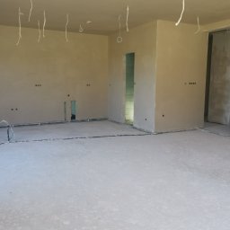 ART-INVEST Damian Solarski - Surowe wnętrze po tynkowaniu maszynowym: widoczne kable wystające z sufitu, gładkie, jasne ściany z otworami na gniazdka elektryczne, oraz wylewka podłogowa z widocznymi przewodami grzewczymi...