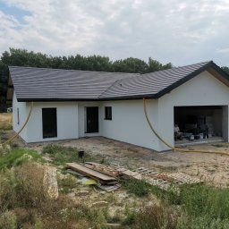 ART-INVEST Damian Solarski - Nowoczesny, parterowy dom z garażem w trakcie prac elewacyjnych, widoczne białe ściany, ciemna dachówka, żółte węże do agregatu tynkarskiego, teren wokół domu częściowo zagospodarowany, w tle las...