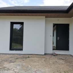 ART-INVEST Damian Solarski - Świeżo otynkowana, biała elewacja domu z czarnymi oknami i drzwiami wejściowymi. Widoczny fragment dachu z ciemną dachówką i podbitką. Na ziemi leży przedłużacz i piasek.