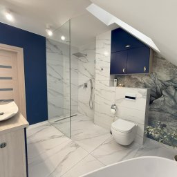 ART-INVEST Damian Solarski - Nowoczesna łazienka na poddaszu z kabiną prysznicową typu walk-in, umywalką nablatową, wiszącą toaletą i szafkami w kolorze granatowym, wykończona białymi płytkami imitującymi marmur i tapetą...