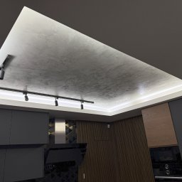 ART-INVEST Damian Solarski - Wnętrze kuchni: Sufit podwieszany z oświetleniem LED i reflektorami szynowymi, wykończony dekoracyjną strukturą przypominającą beton architektoniczny, na tle zabudowy kuchennej w odcieniach...
