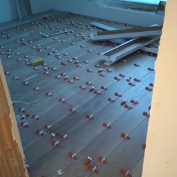 ART-INVEST Damian Solarski - Układanie szarych płytek podłogowych z użyciem czerwonych i białych klinów dystansowych oraz żółtej gąbki, widok z góry na niedokończoną podłogę w nowym budynku.