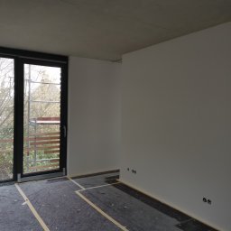 ART-INVEST Damian Solarski - Wnętrze pokoju w stanie deweloperskim po wykonaniu gładzi, z zabezpieczoną podłogą, widoczne puszki elektryczne i okno z widokiem na rusztowanie.