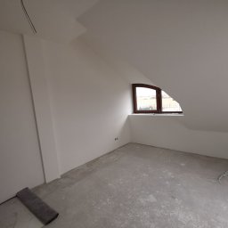 ART-INVEST Damian Solarski - Poddasze po wykonaniu gładzi, widoczne białe ściany, okno w drewnianej ramie, szary betonowy podkład i zwinięty szary dywanik przy ścianie.