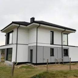 Dwukondygnacyjny dom jednorodzinny z jasną elewacją, ciemną stolarką okienną i grafitowym wykończeniem dolnej części fasady, widoczny z zewnątrz przez ogrodzenie z siatki.