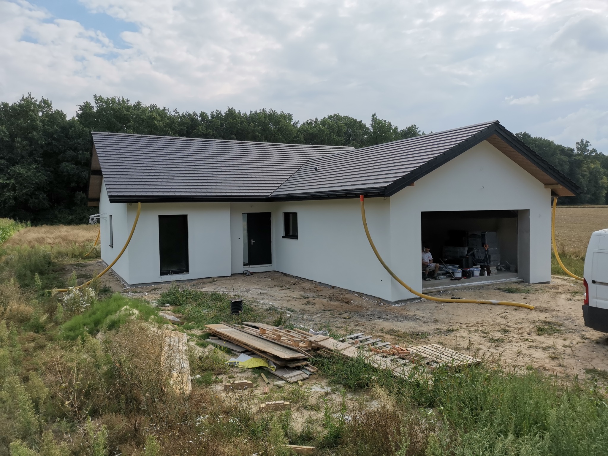 Nowoczesny, parterowy dom z garażem w trakcie prac elewacyjnych, widoczne białe ściany, ciemna dachówka, żółte węże do agregatu tynkarskiego, teren wokół domu częściowo zagospodarowany, w tle las...