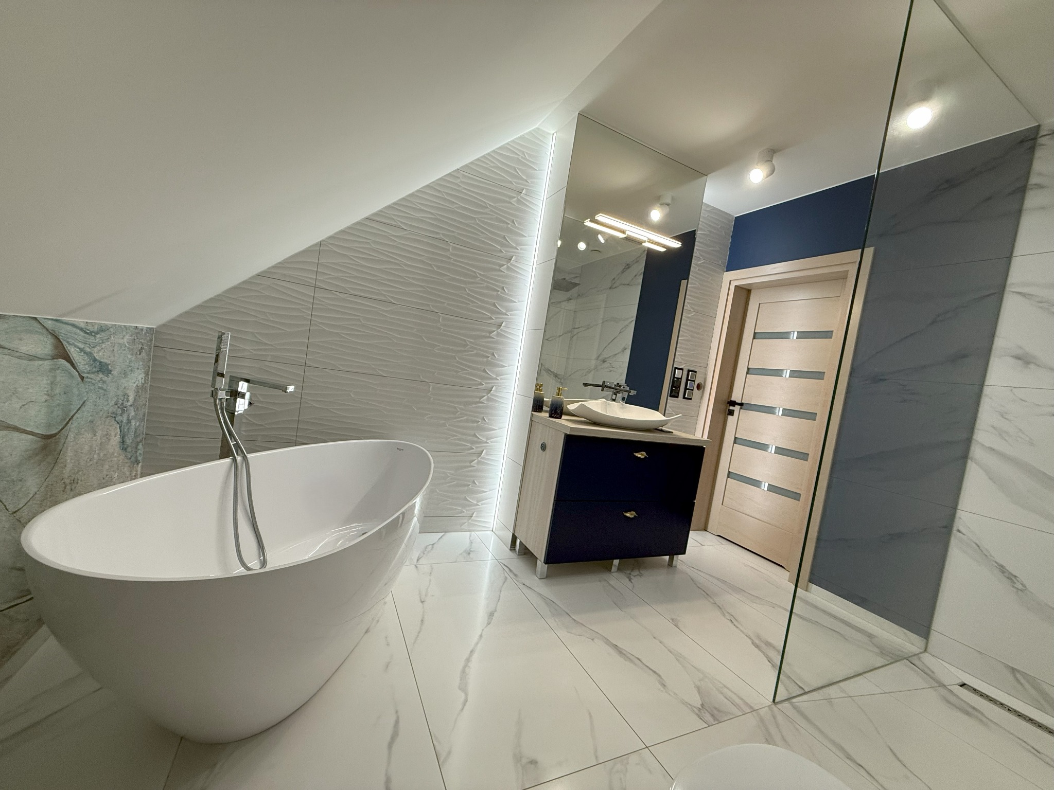 Nowoczesna łazienka na poddaszu w Puchaczowie z wolnostojącą białą wanną, umywalką nablatową na szafce w kolorze granatu i beżu, oraz szklaną kabiną prysznicową typu walk-in.