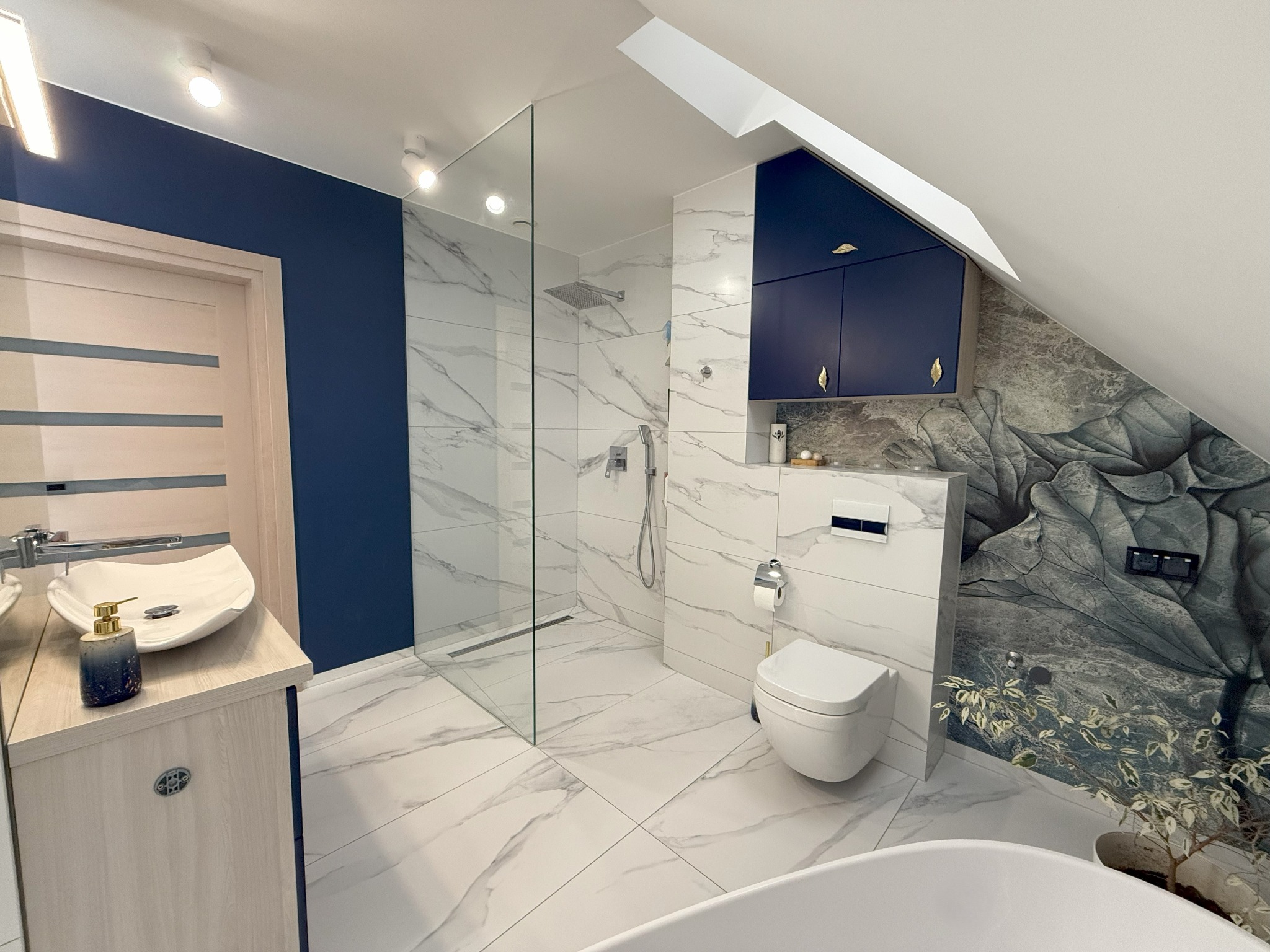 Nowoczesna łazienka na poddaszu z kabiną prysznicową typu walk-in, umywalką nablatową, wiszącą toaletą i szafkami w kolorze granatowym, wykończona białymi płytkami imitującymi marmur i tapetą...