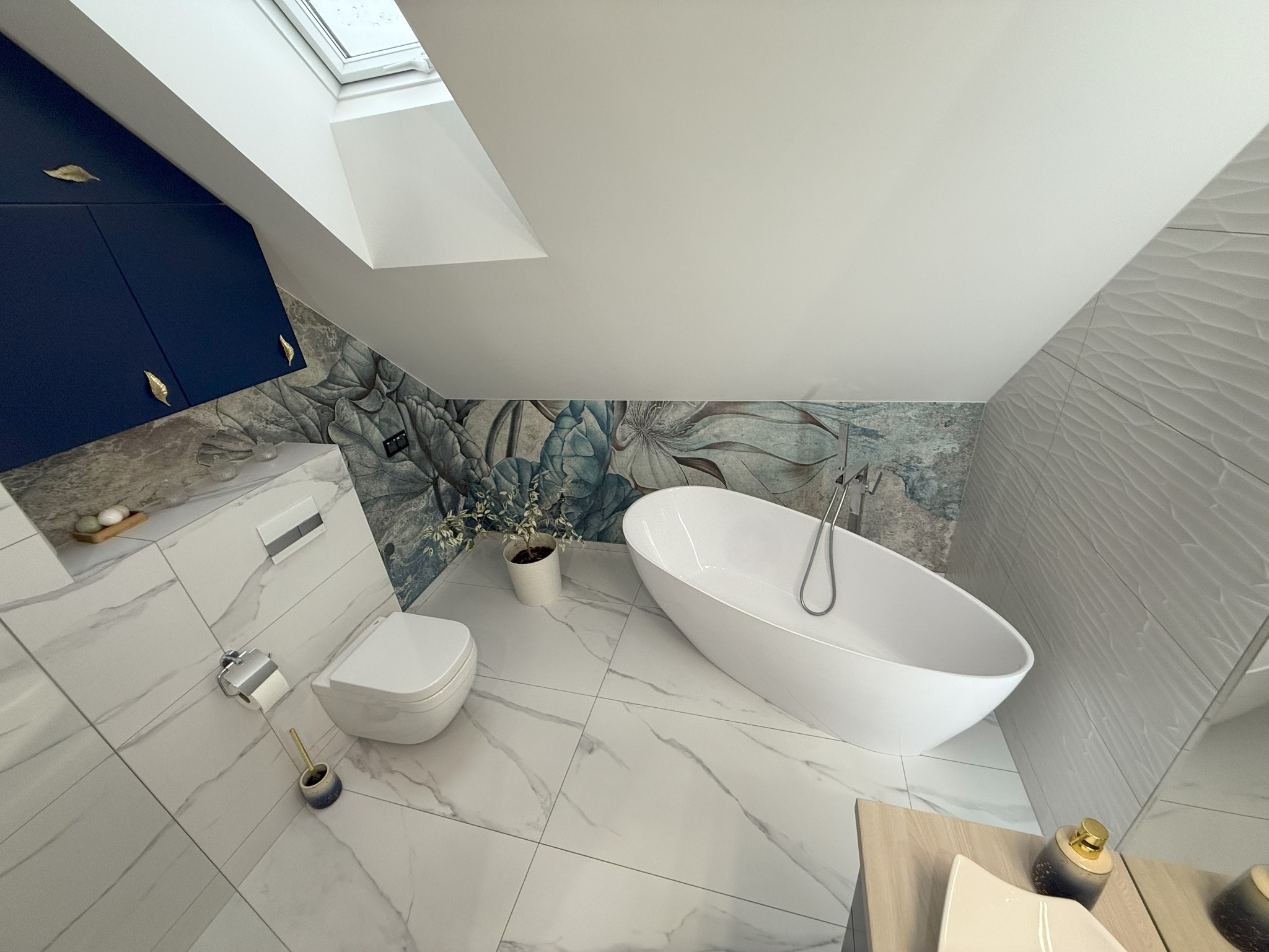 Nowoczesna łazienka na poddaszu ze skośnym sufitem, białą wolnostojącą wanną, toaletą, marmurową podłogą i ścianą z motywem kwiatowym, oświetlona oknem dachowym.