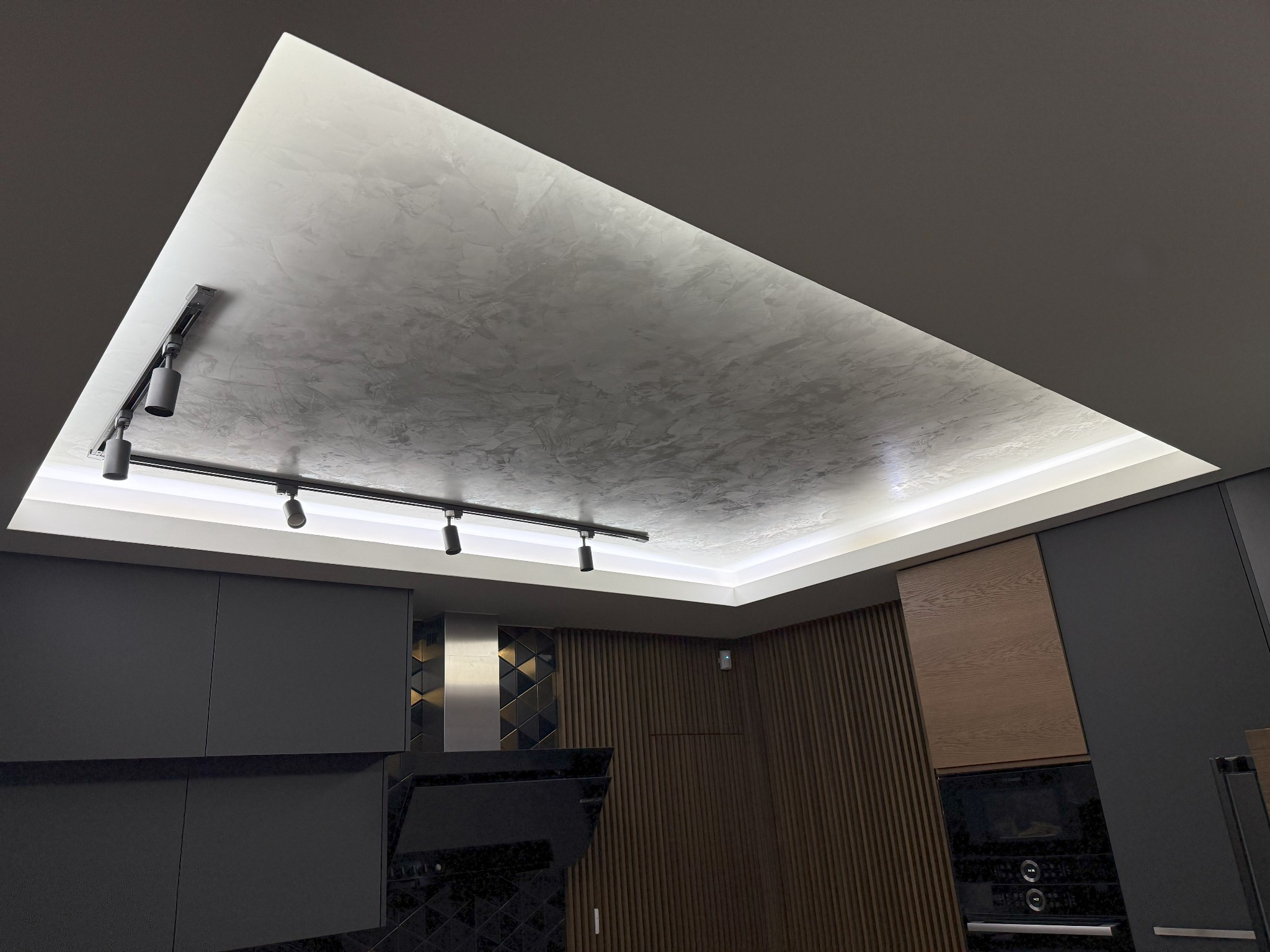 Wnętrze kuchni: Sufit podwieszany z oświetleniem LED i reflektorami szynowymi, wykończony dekoracyjną strukturą przypominającą beton architektoniczny, na tle zabudowy kuchennej w odcieniach...