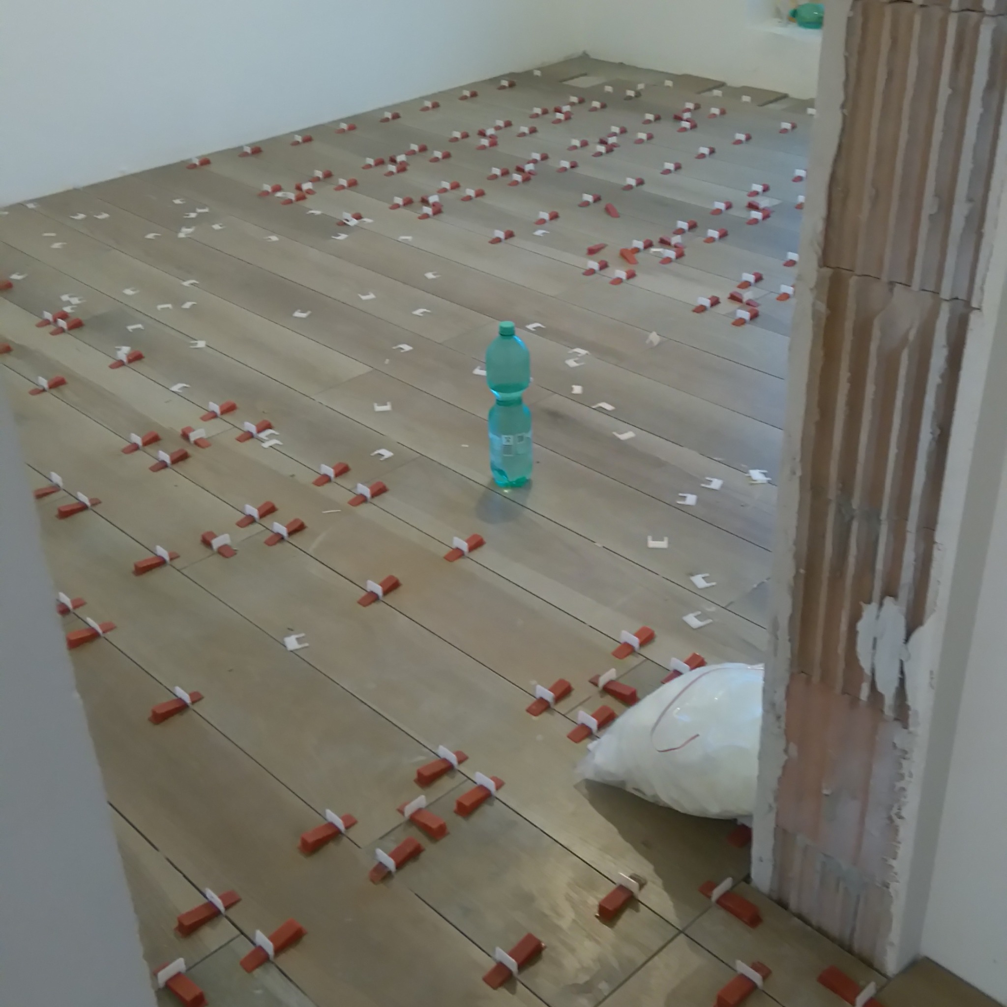 Układanie podłogi z płytek imitujących drewno, z użyciem czerwonych klinów i białych krzyżyków dystansowych. W tle butelka wody i worek z zaprawą.