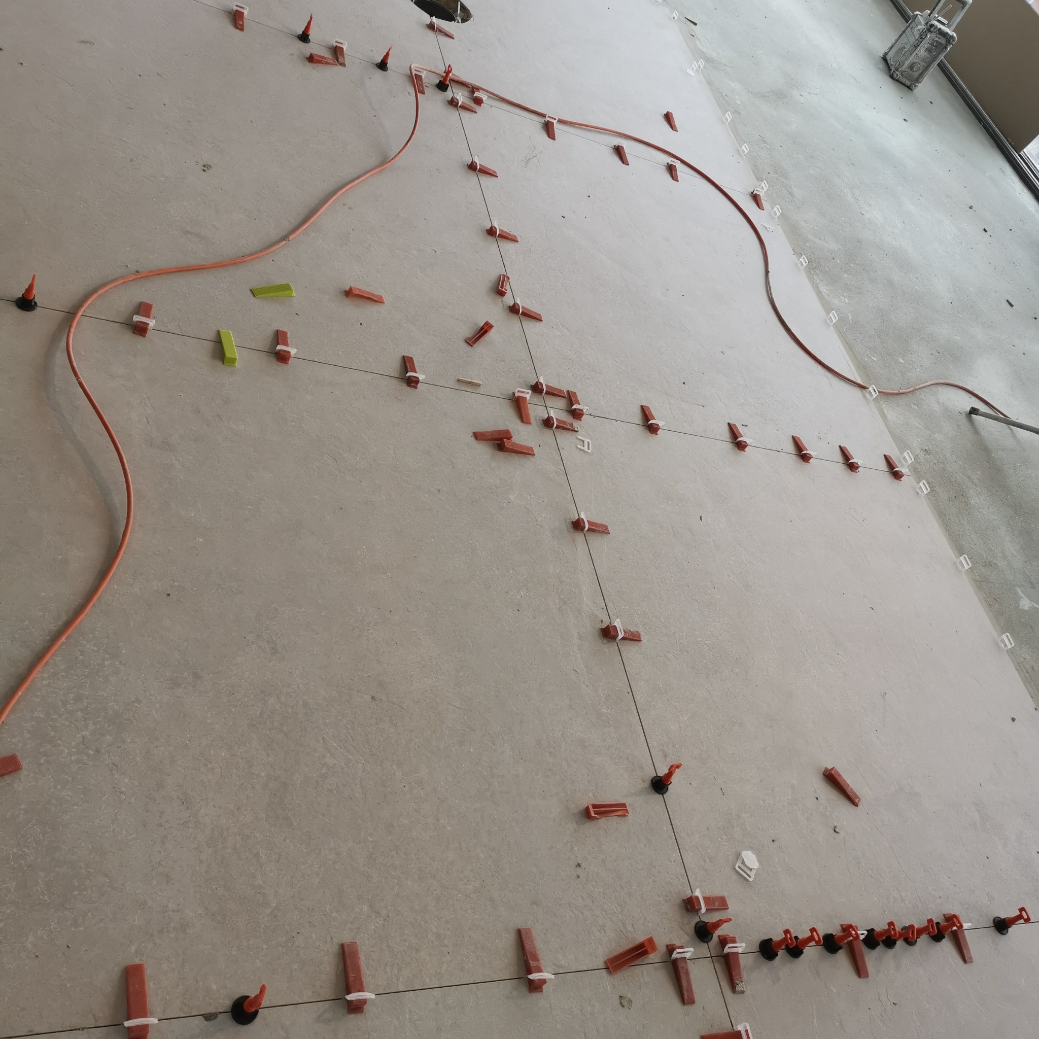 Układanie płytek podłogowych z wykorzystaniem systemu poziomowania, widoczne krzyżyki dystansowe, kliny i zaciski, pomarańczowy przewód elektryczny na szarej powierzchni.