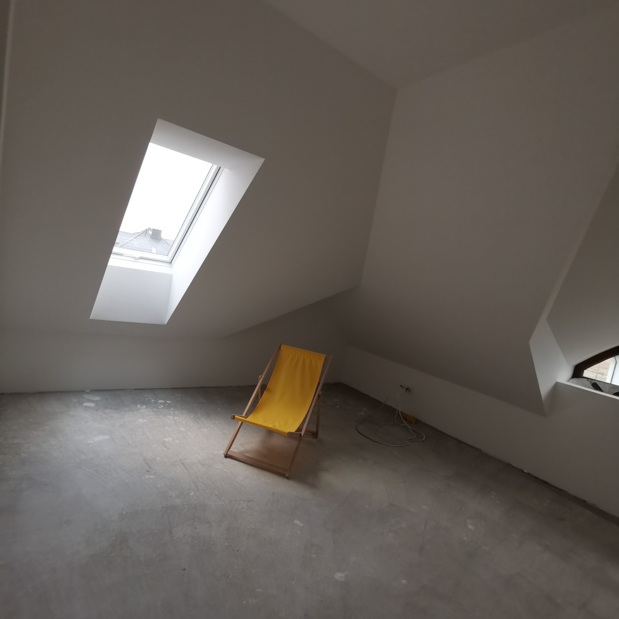 Poddasze po remoncie z żółtym leżakiem na środku, widoczne okno dachowe i świeżo położone gładzie na ścianach.