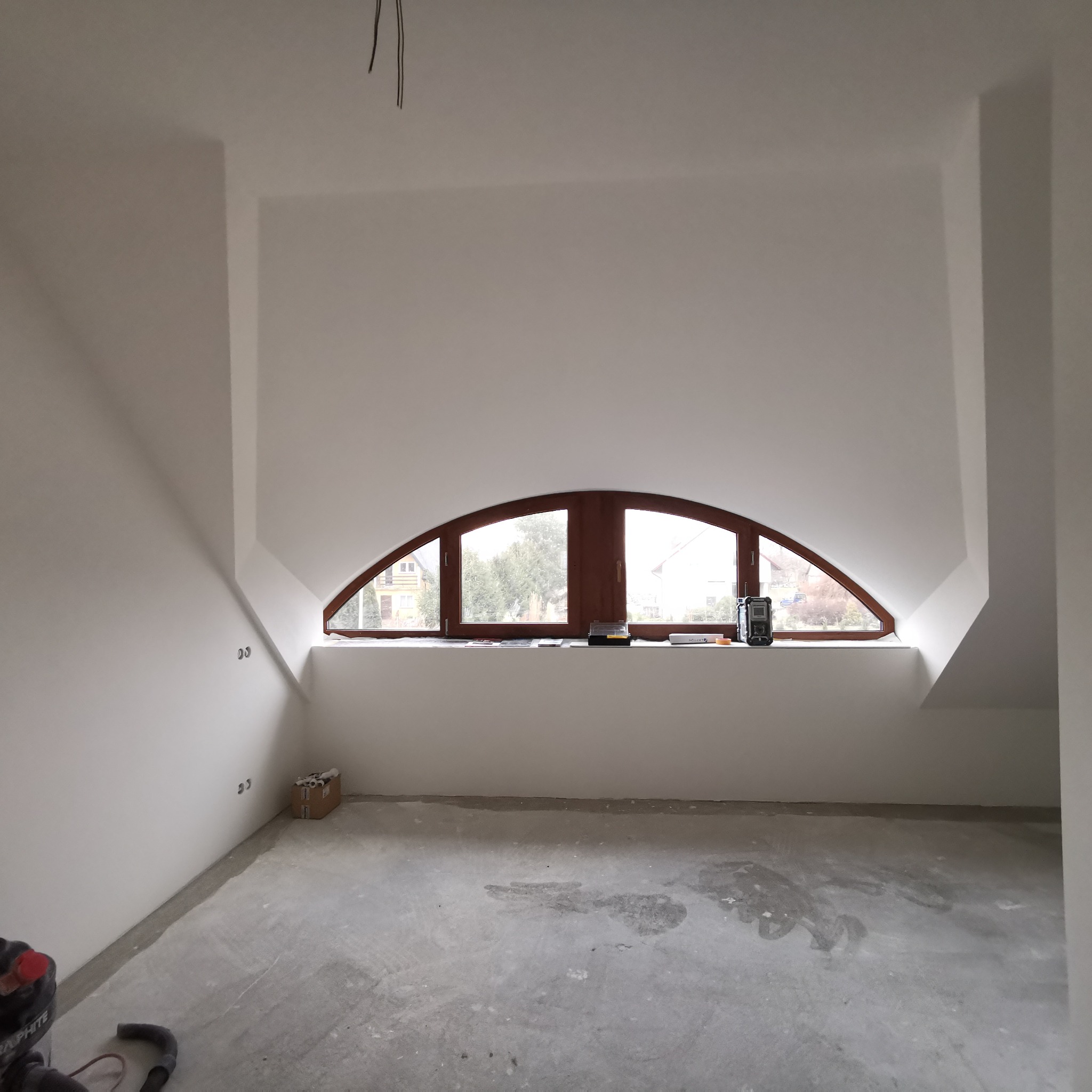 Poddasze z oknem w kształcie łuku po wykonaniu gładzi, widoczny szary betonowy podkład podłogowy i wystające przewody elektryczne.