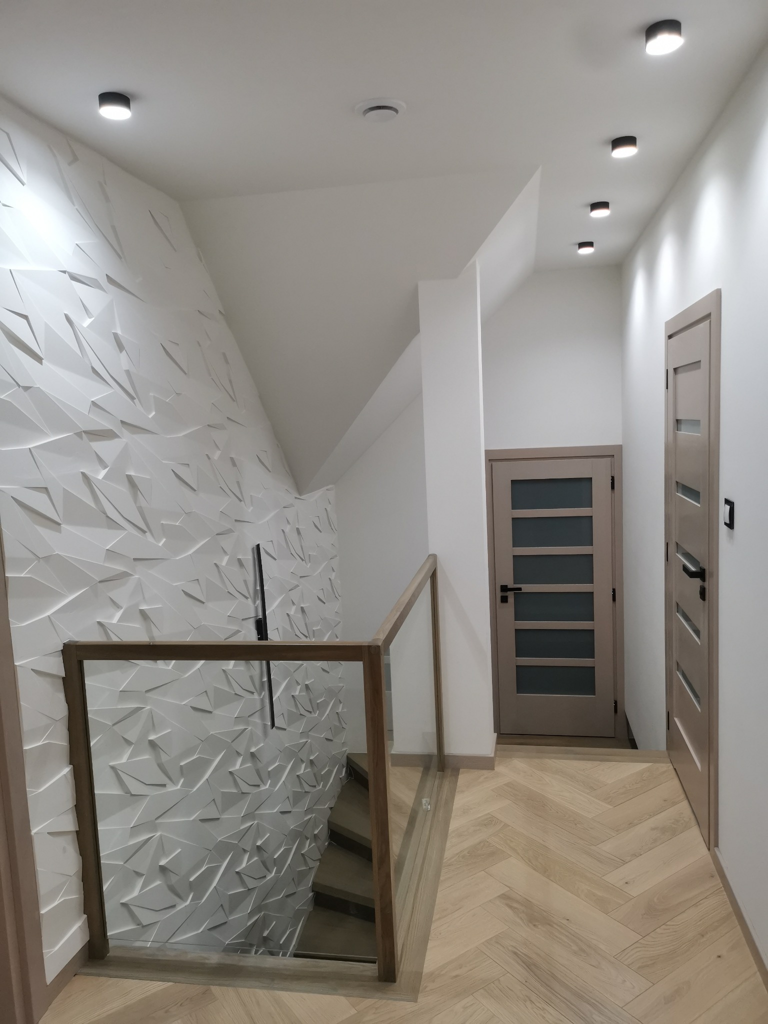 Nowoczesne wnętrze z trójwymiarową białą ścianą, drewnianą balustradą schodów ze szkłem, parkietem w jodełkę i dwiema beżowymi parami drzwi.