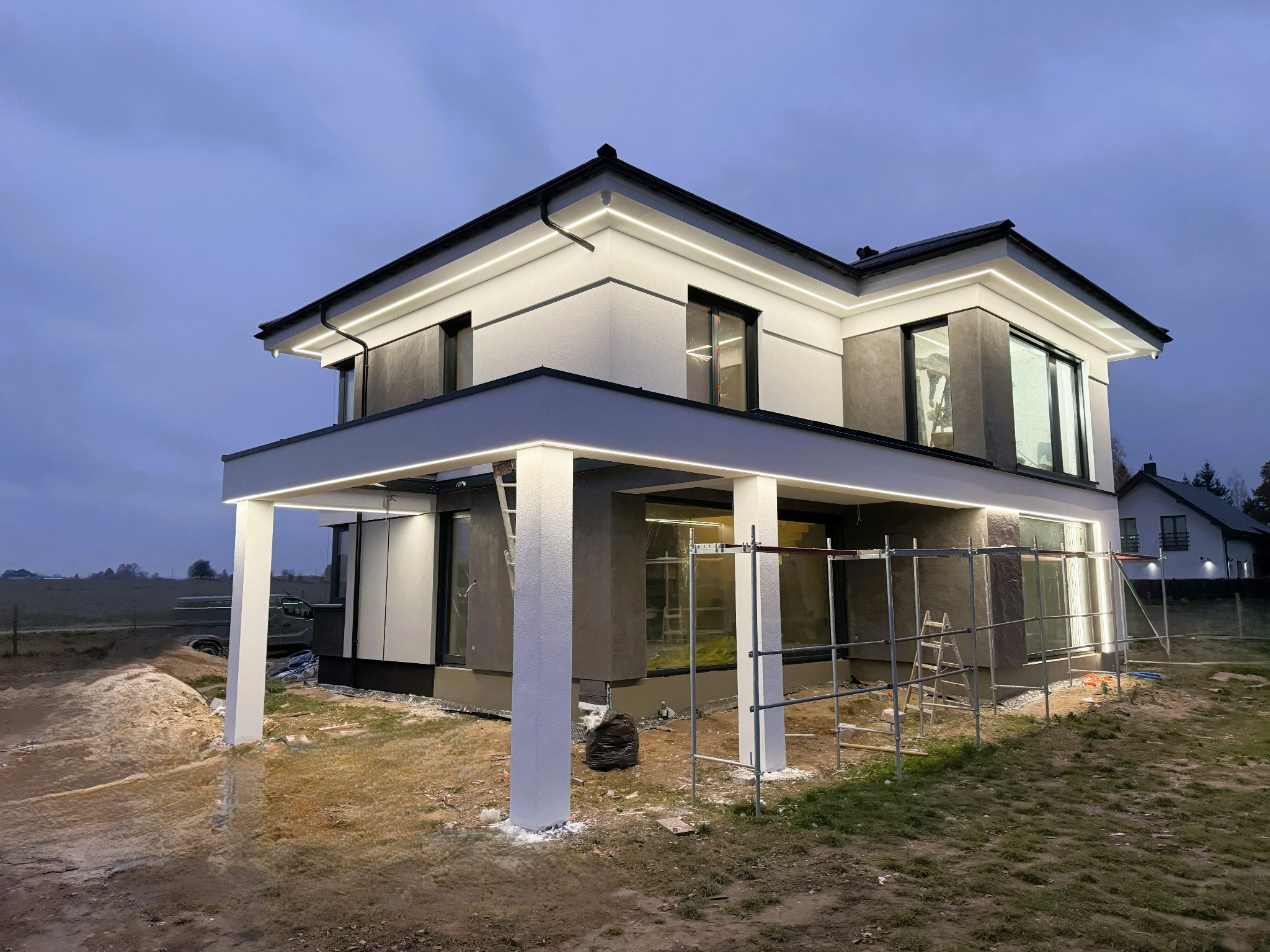 Dwukondygnacyjny dom z płaskim dachem w trakcie prac elewacyjnych, widoczne rusztowania, oświetlenie LED na elewacji, teren wokół domu niezagospodarowany.