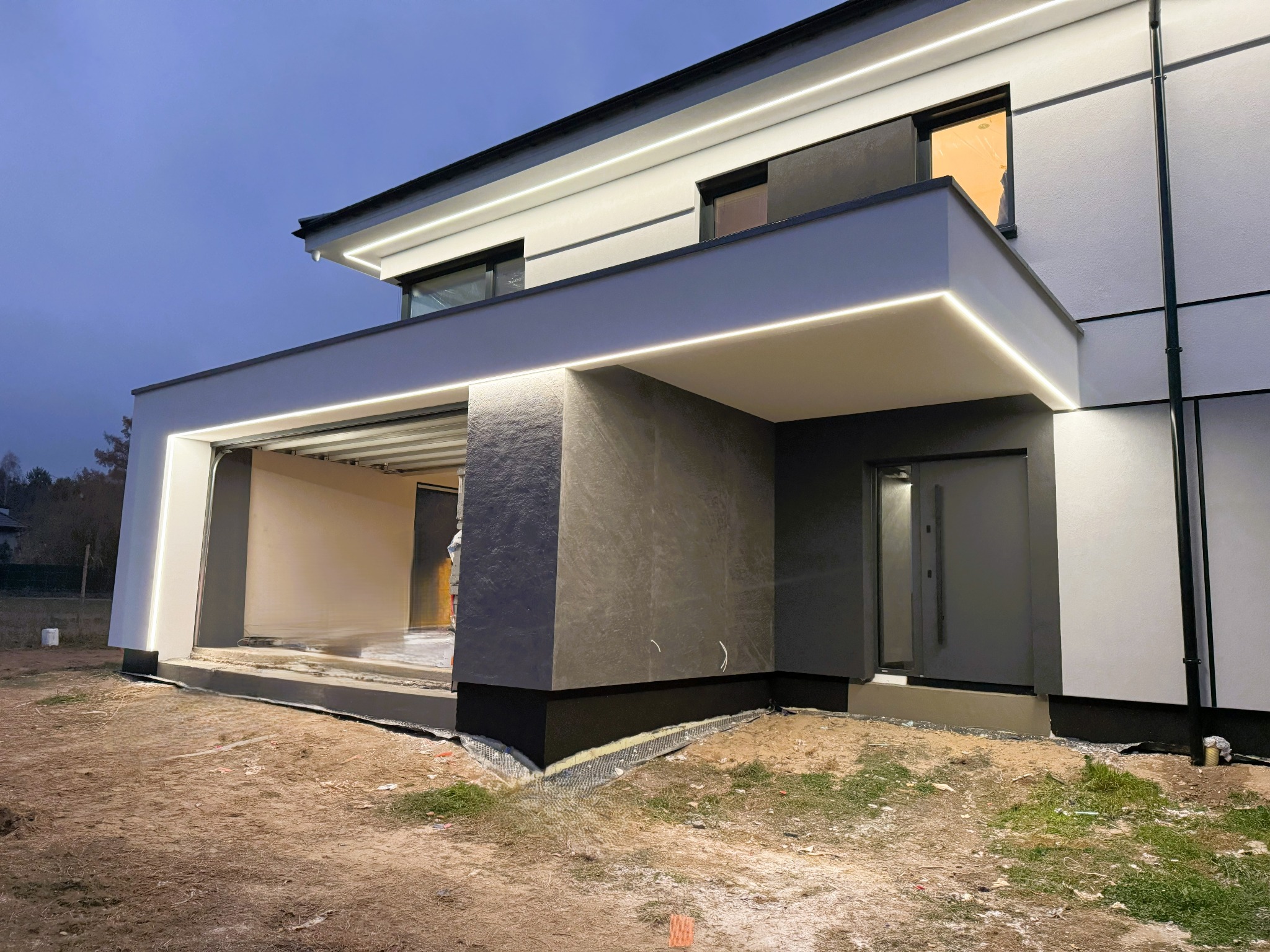 Nowoczesny dom jednorodzinny z oświetleniem LED elewacji, widok z zewnątrz na częściowo wykończoną elewację i teren przed budynkiem.