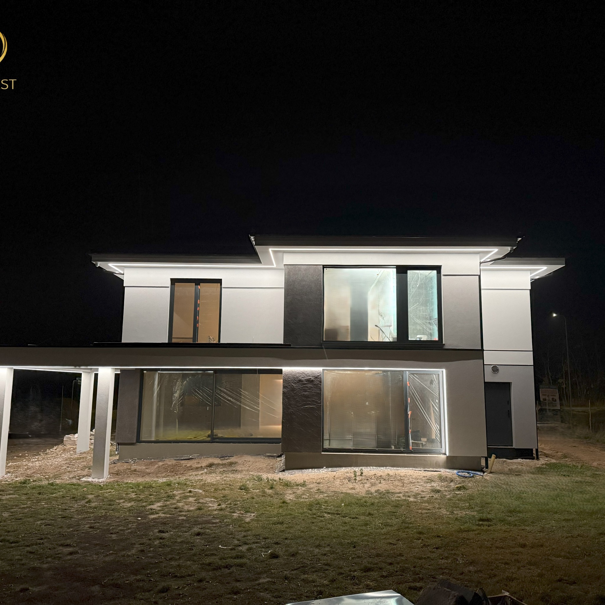 Nowoczesny dom jednorodzinny w trakcie wykańczania elewacji, widoczny nocą z podświetleniem LED, okna zabezpieczone folią, teren wokół domu częściowo zagospodarowany.