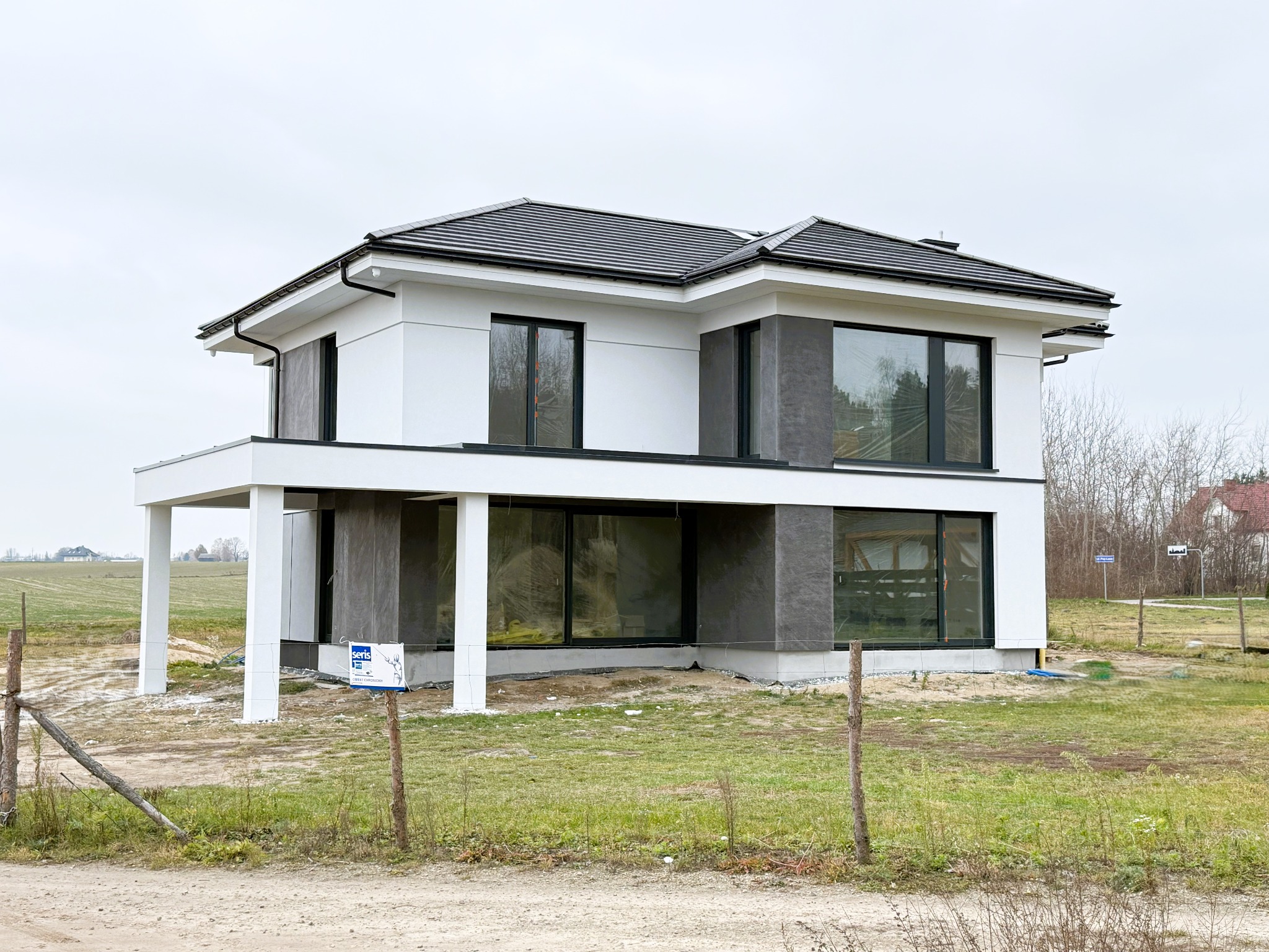 Dwupiętrowy dom w stylu nowoczesnym z płaskim dachem i dużymi oknami, elewacja częściowo biała, częściowo wykończona szarym kamieniem dekoracyjnym, teren wokół domu jeszcze niezagospodarowany...