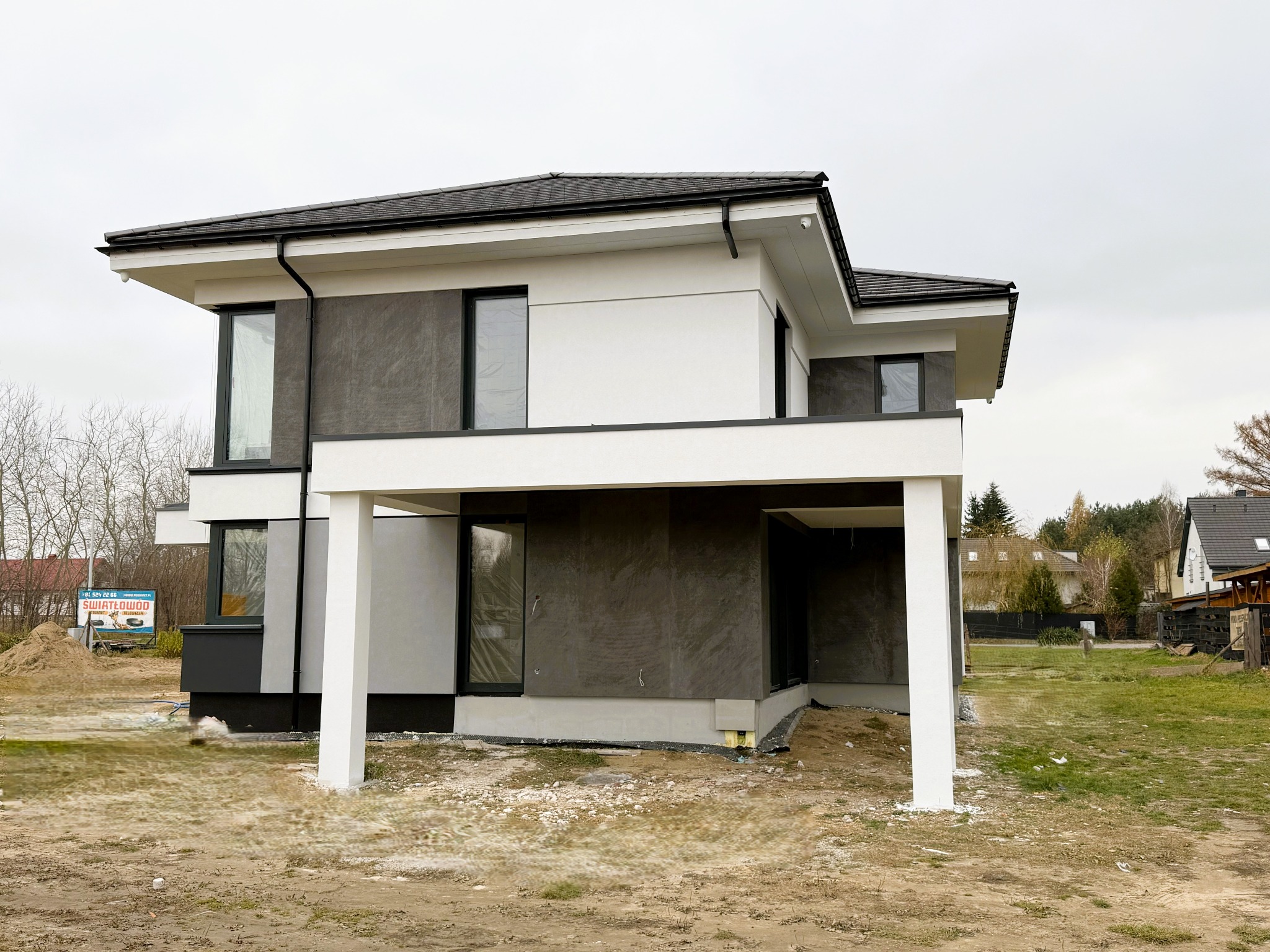 Nowoczesny dom jednorodzinny z elewacją w kolorze białym i szarym, widoczne elementy konstrukcyjne podtrzymujące balkon, teren wokół domu niezagospodarowany, widoczny billboard w tle.