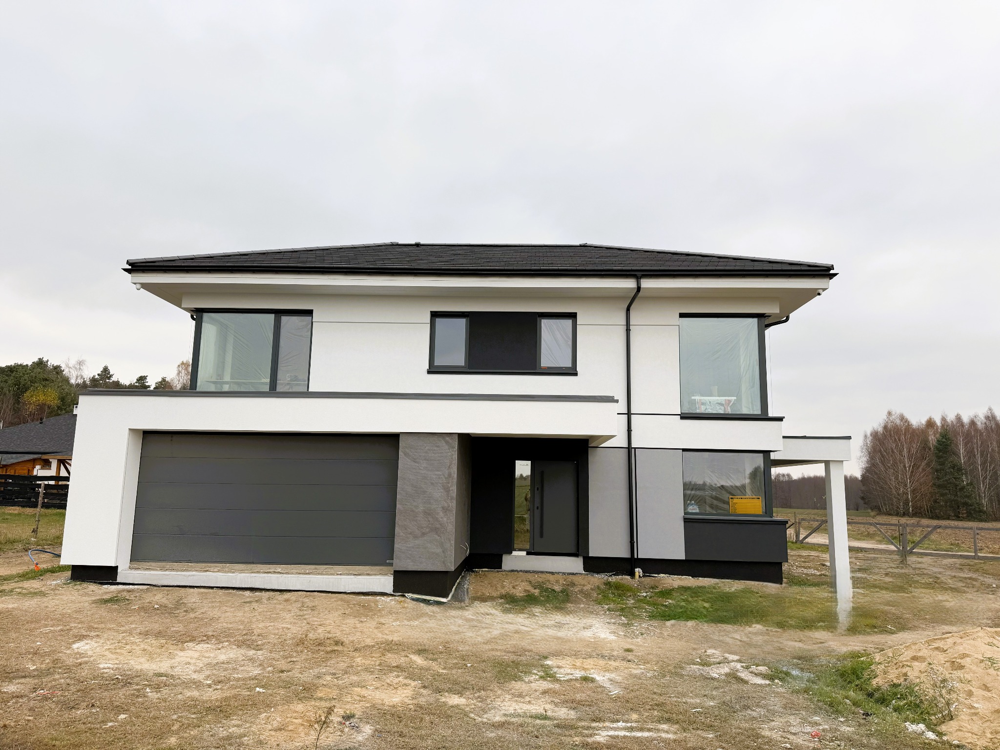 Nowoczesny dom jednorodzinny z płaskim dachem, białą elewacją i grafitową bramą garażową oraz stolarką okienną, widoczny w surowym krajobrazie na etapie wykończenia.