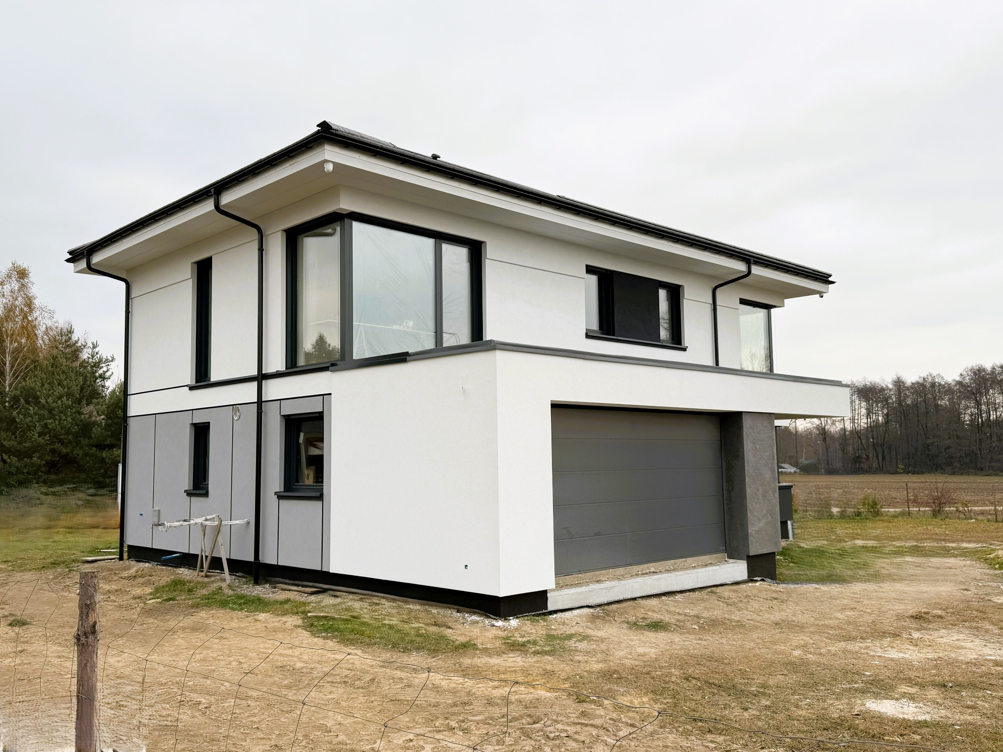 Nowoczesny, dwukondygnacyjny dom z szarą elewacją na parterze i białą na piętrze, z garażem wbudowanym, widoczne rusztowanie przy ścianie bocznej, teren wokół domu jeszcze niezagospodarowany.