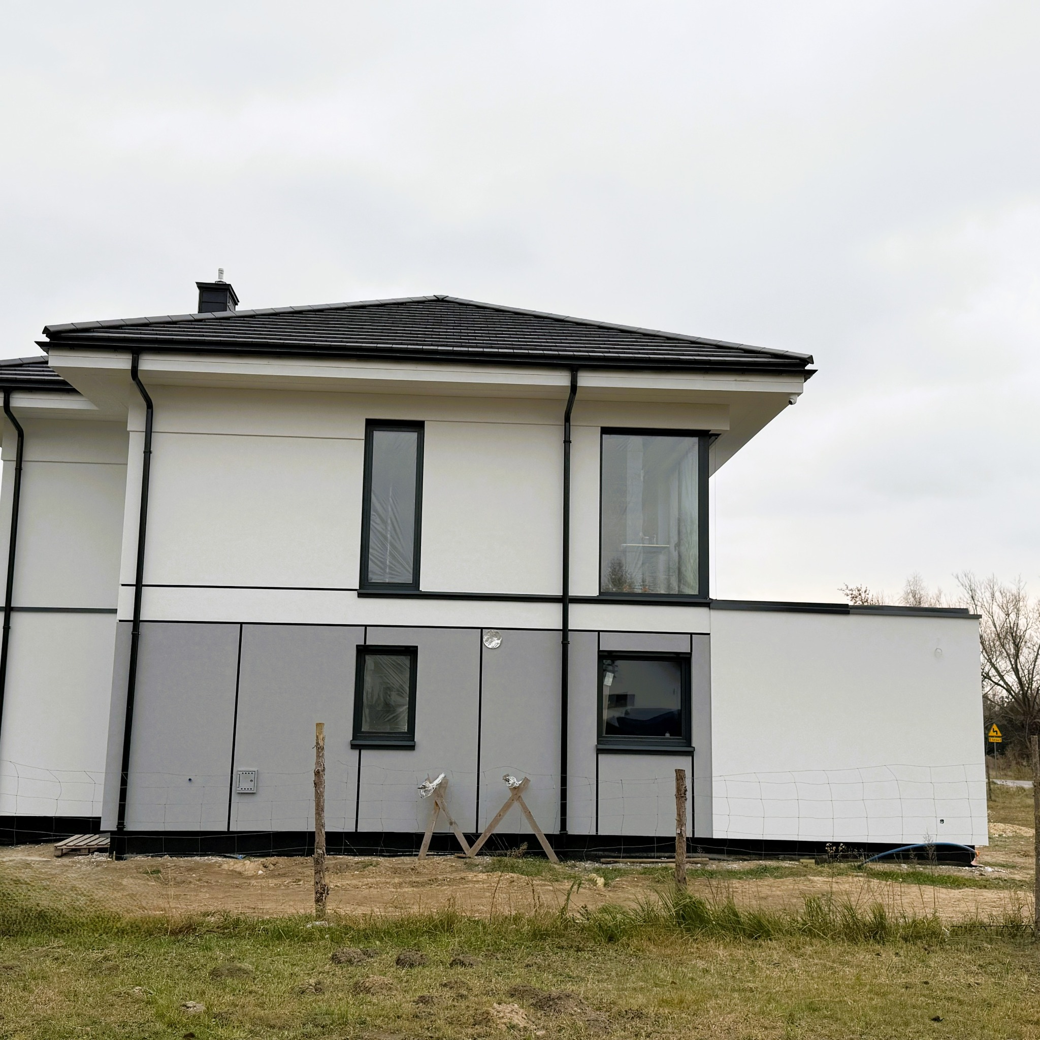 Nowoczesny dom jednorodzinny z biało-szarą elewacją i czarnymi oknami, widoczny z zewnątrz, z fragmentem trawnika i ogrodzenia z siatki.