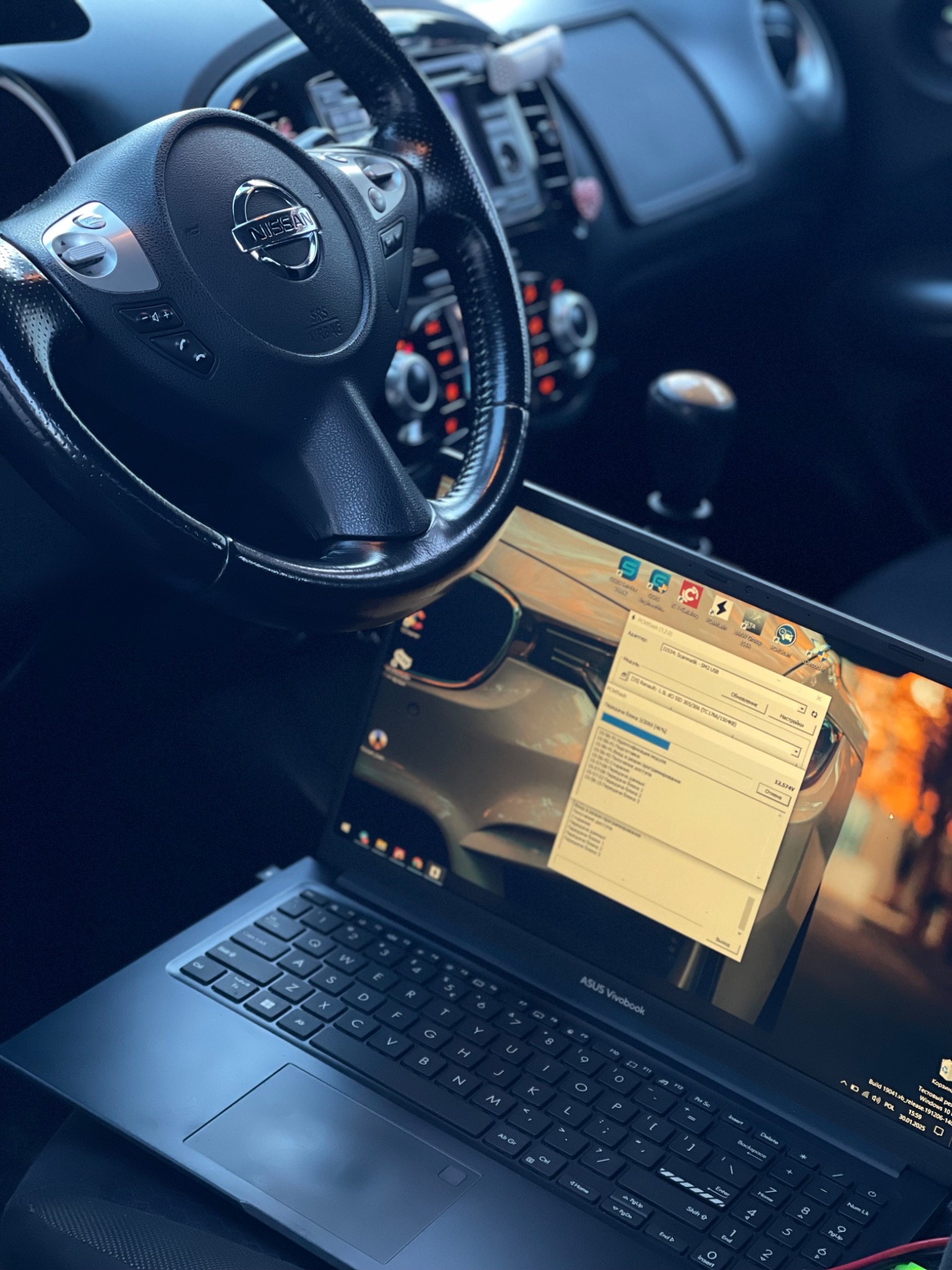 Wnętrze samochodu Nissan z laptopem ASUS Vivobook na siedzeniu, na ekranie widoczne oprogramowanie diagnostyczne z paskiem postępu, kierownica i deska rozdzielcza w tle.