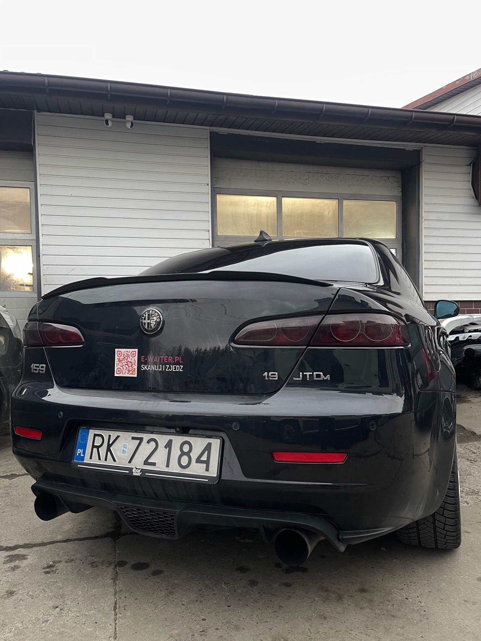 Tył czarnej Alfy Romeo 159 JTDm zaparkowanej przed budynkiem z garażem; widoczny spoiler, naklejka z kodem QR, polska tablica rejestracyjna i podwójny wydech.