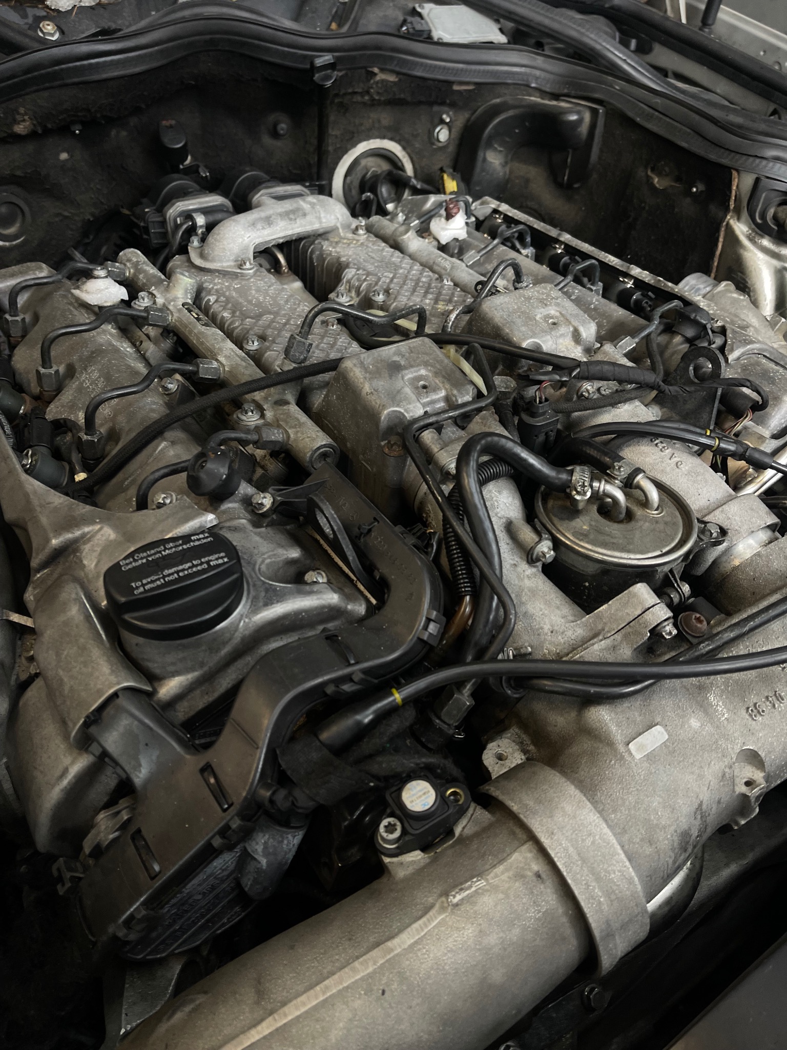 Widok z bliska na silnik samochodowy podczas naprawy, widoczne przewody, elementy metalowe i plastikowe, ślady użytkowania i zabrudzenia, czarny korek wlewu oleju z napisem.