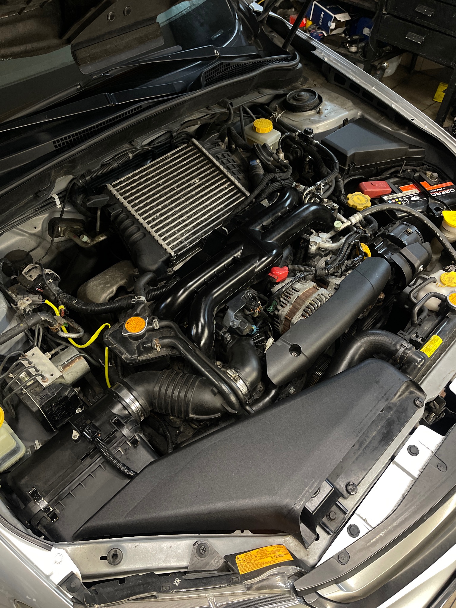 Widok z góry na otwartą maskę srebrnego samochodu z widocznym silnikiem, akumulatorem i innymi komponentami, z żółtymi akcentami na korkach i przewodach.