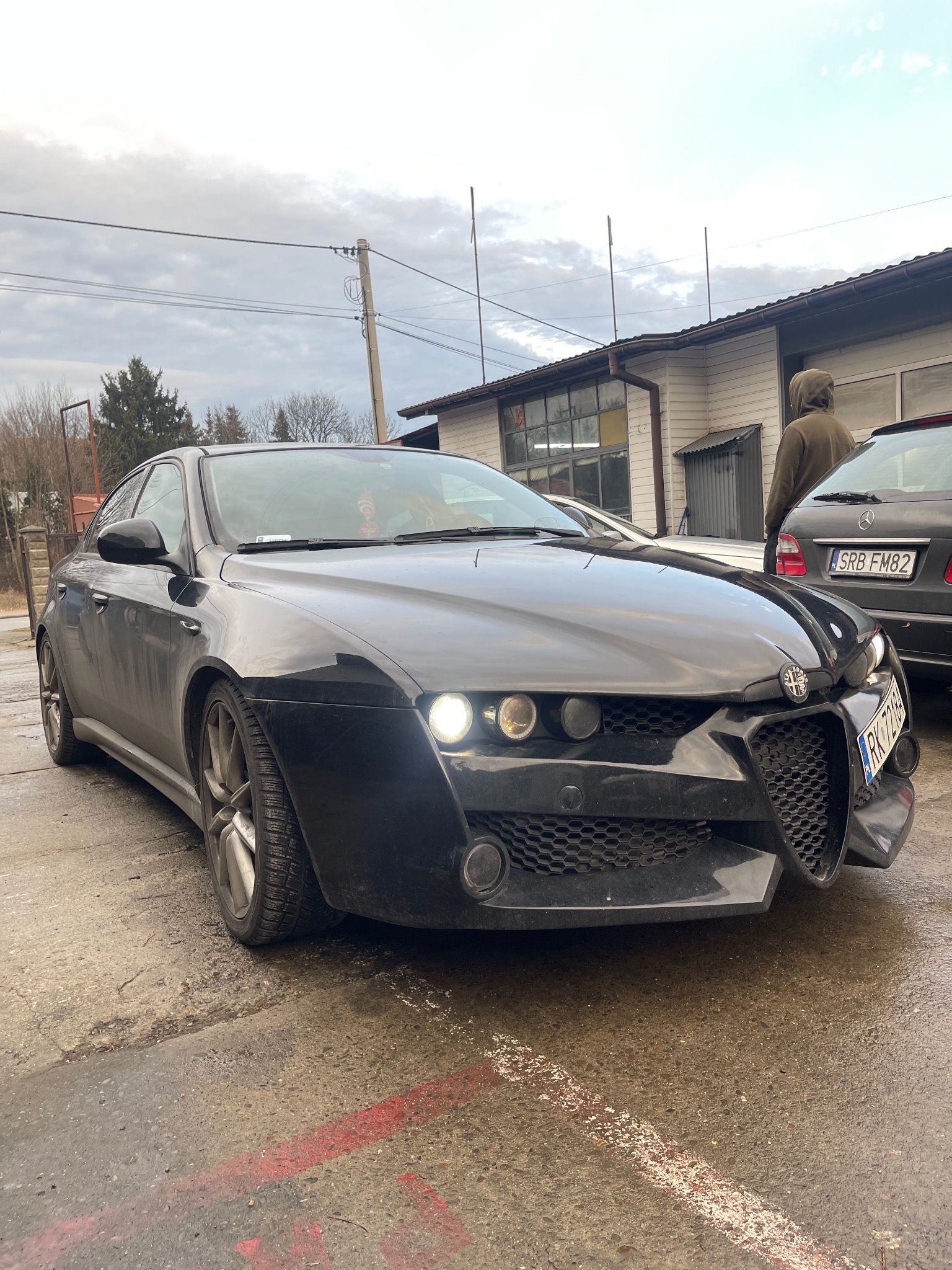 Ciemny, lekko zabrudzony samochód Alfa Romeo stoi na mokrym betonowym placu przed budynkiem warsztatu samochodowego; w tle widoczny inny samochód i osoba.