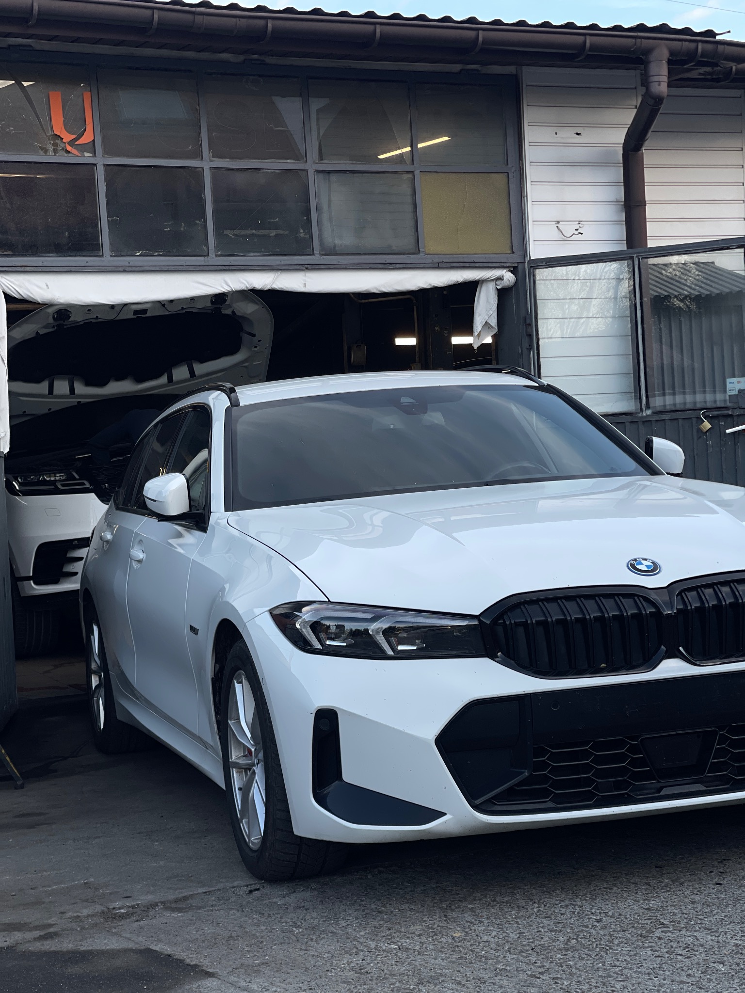 Białe BMW serii 3 stoi przed warsztatem z otwartą maską innego białego SUV-a w tle, widoczne logo BMW na masce.
