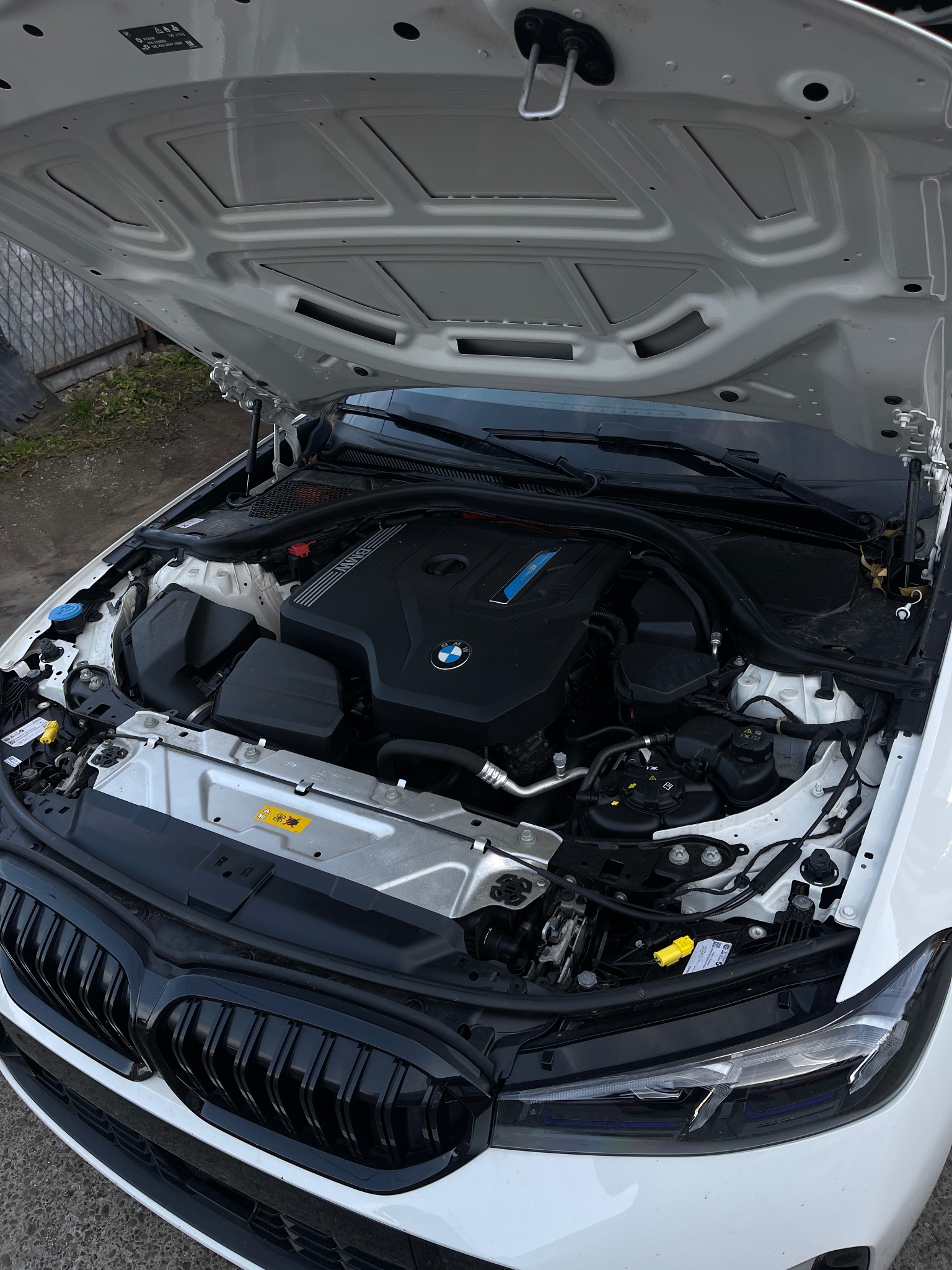 Otwarta maska białego samochodu BMW, widoczny silnik z elementami w kolorze czarnym i srebrnym, detale układu chłodzenia i przewodów, oraz czarny grill z przodu pojazdu.