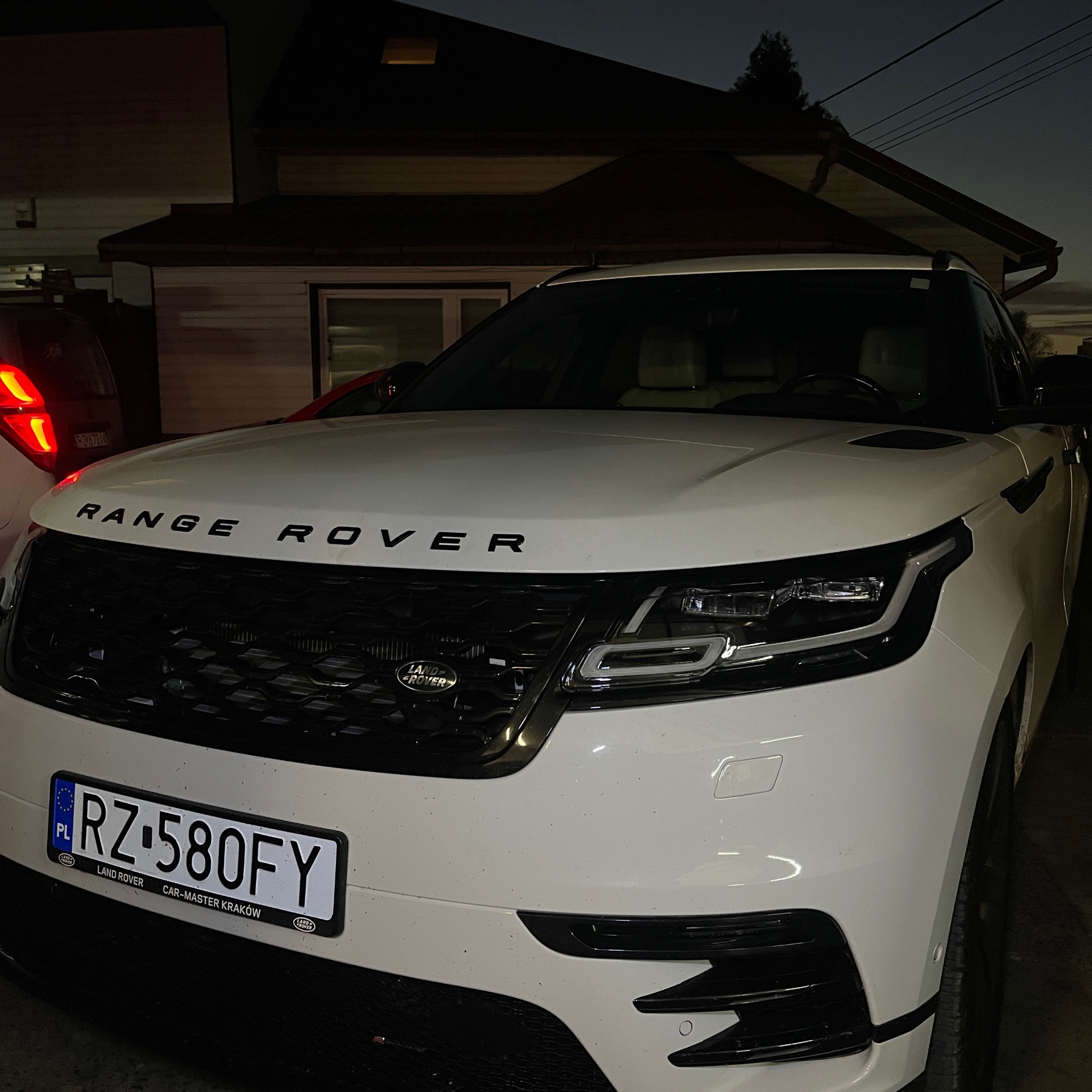 Biały Range Rover Velar z polską tablicą rejestracyjną RZ 580FY, zaparkowany przed domem w słabym oświetleniu wieczornym, widoczne zabrudzenia na karoserii.