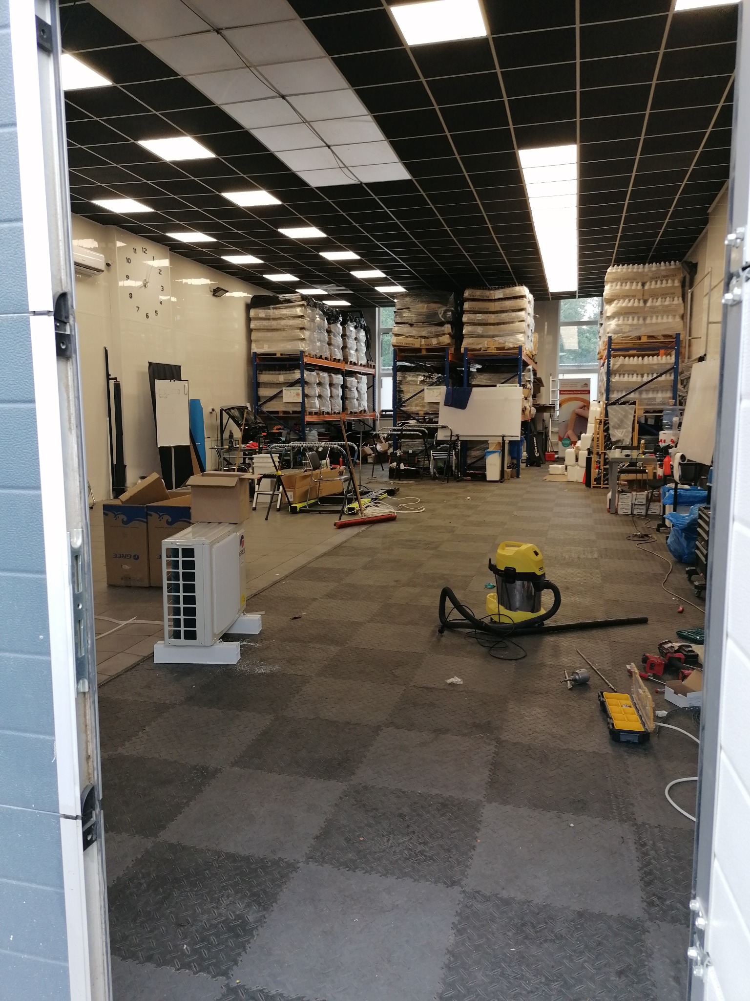 Wnętrze magazynu z klimatyzatorem Gree na podłodze, widok z otwartych drzwi, narzędzia i odkurzacz widoczne w tle.