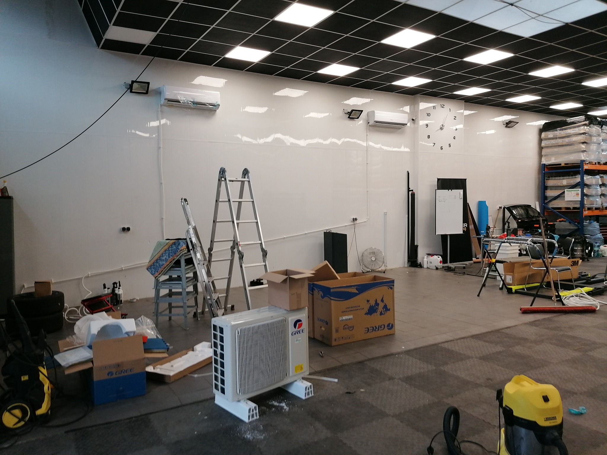 Przygotowania do montażu klimatyzacji w biurze: jednostka zewnętrzna marki Gree, drabiny, kartony i narzędzia na podłodze, widoczne zainstalowane jednostki wewnętrzne na ścianie. Białe ściany...