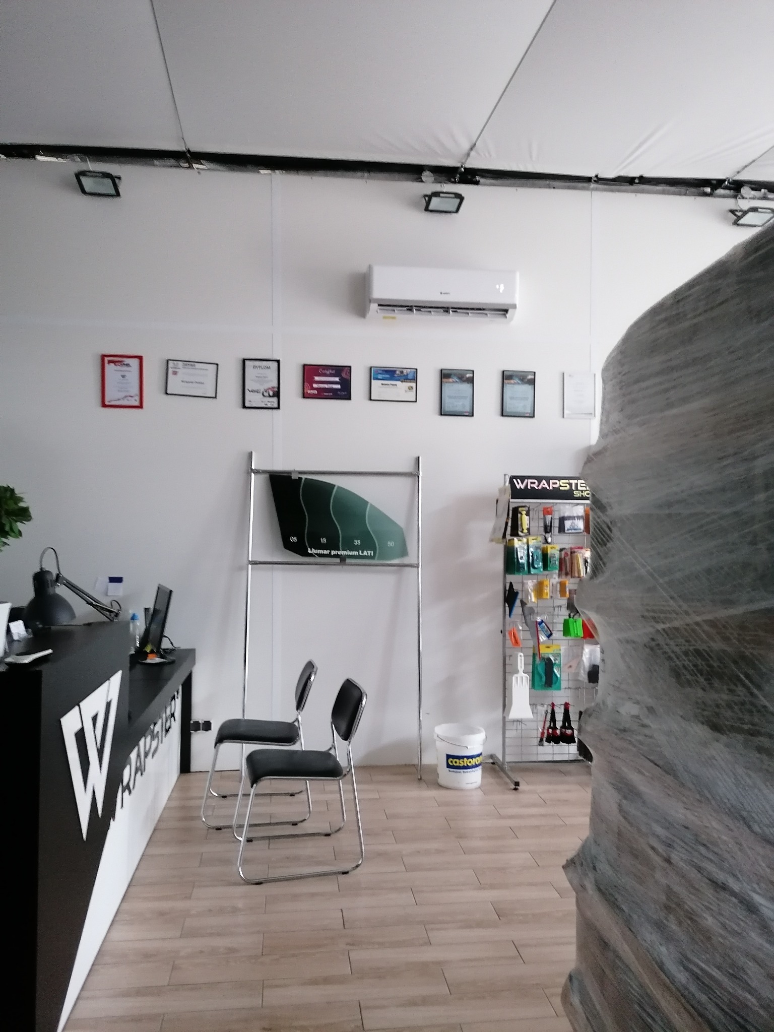 Wnętrze biura firmy Wrapster z klimatyzatorem na ścianie, certyfikatami, wzornikiem folii okiennych, regałem z akcesoriami i owiniętym folią autem na pierwszym planie.
