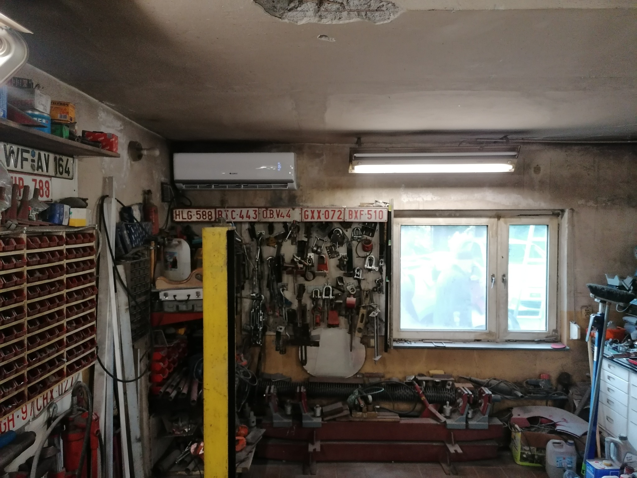 Wnętrze warsztatu z zamontowanym klimatyzatorem na ścianie, pod nim tablica z narzędziami i kolekcją tablic rejestracyjnych, widoczne regały z narzędziami i akcesoriami